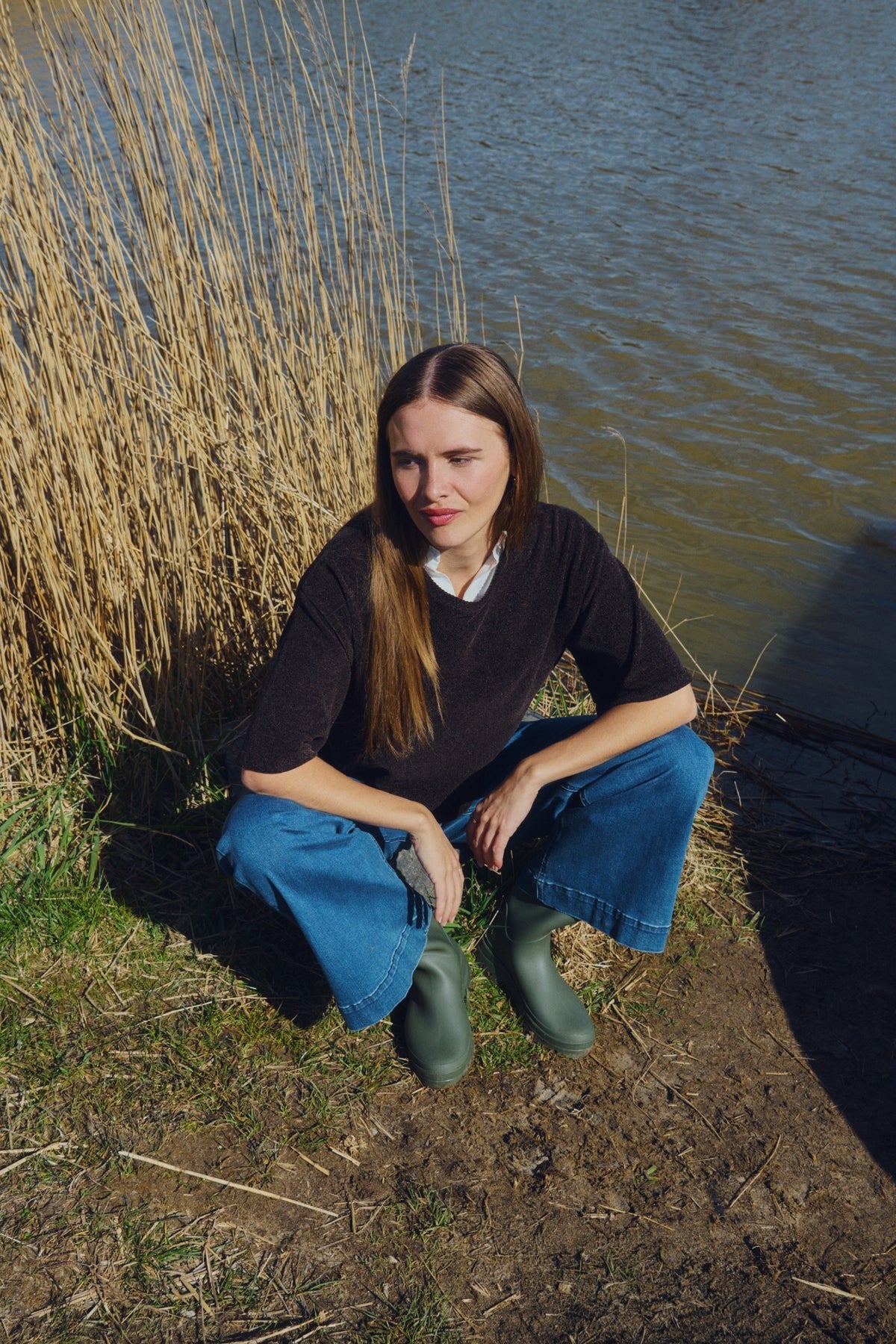 En kvinde iført en Freequent Tempo-bluse i mørkebrun, jeans og grønne støvler sidder på hug ved en dam med tørt græs på en solskinsdag.
