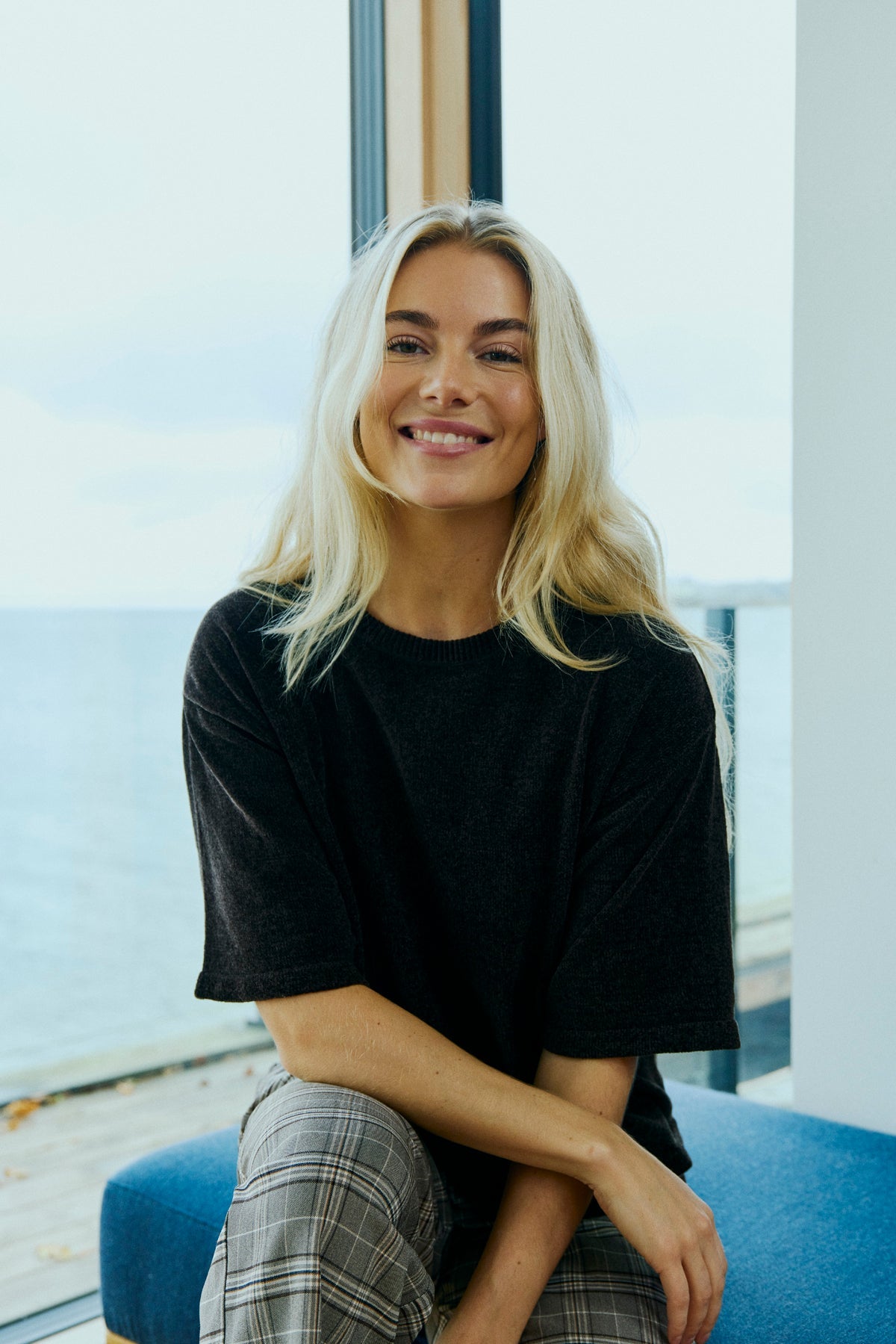 En kvinde med langt blondt hår smiler ved et vindue, iført en Freequent Tempo Mørkebrun striktrøje og ternede bukser.
