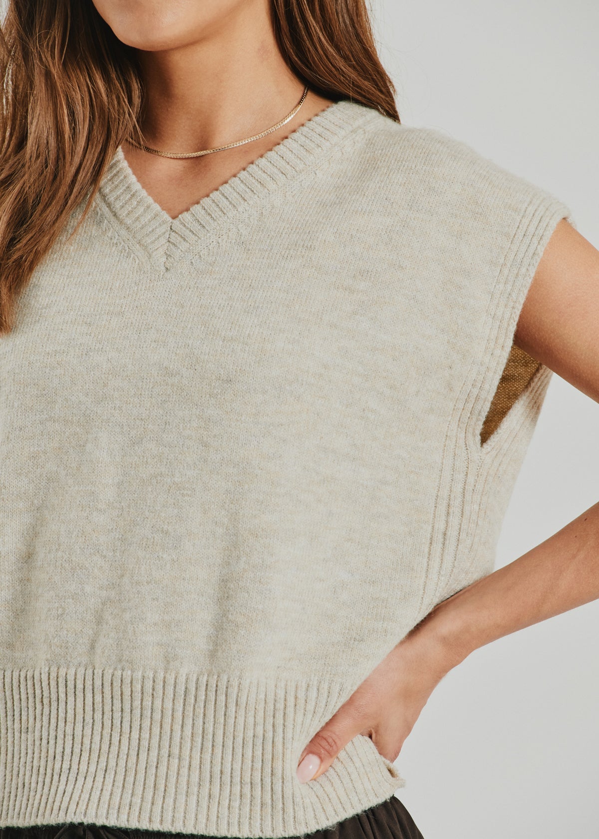 A woman stands with one hand on her hip, wearing the Freequent Dustin Beige strikvest (155348) in a soft wool blend. The vest features a light beige color, V-neckline, and ribbed hem. Her long brown hair is worn down, complemented by a simple necklace.