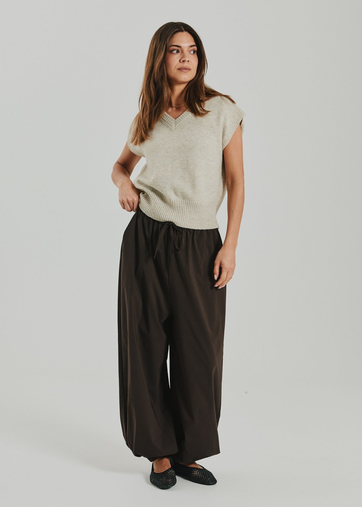 A woman with long brown hair stands against a plain light background, wearing the Freequent Dustin Beige sleeveless V-neck knit vest, paired with loose dark brown wide-leg pants and black shoes.