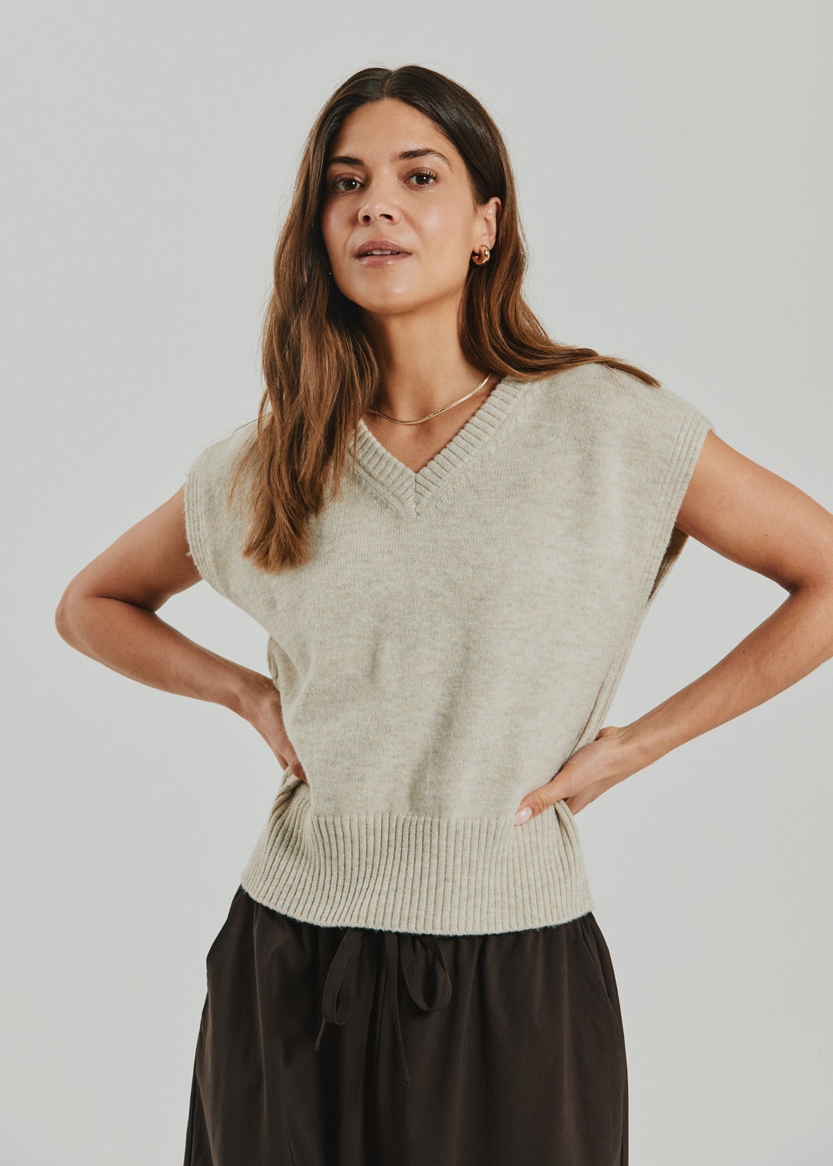 A woman with long brown hair wears the Freequent - Dustin Beige 155348 knit vest in an uldblanding, paired with dark pants, standing with her hands on her hips against a plain light background.