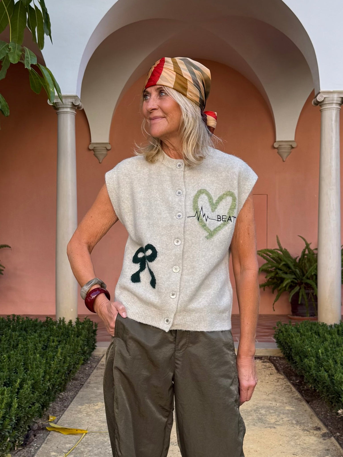 En kvinde med tørklæde og en beige vest fra Freequent Nomas står i en gårdhave med grønne planter og buegange bag sig.
