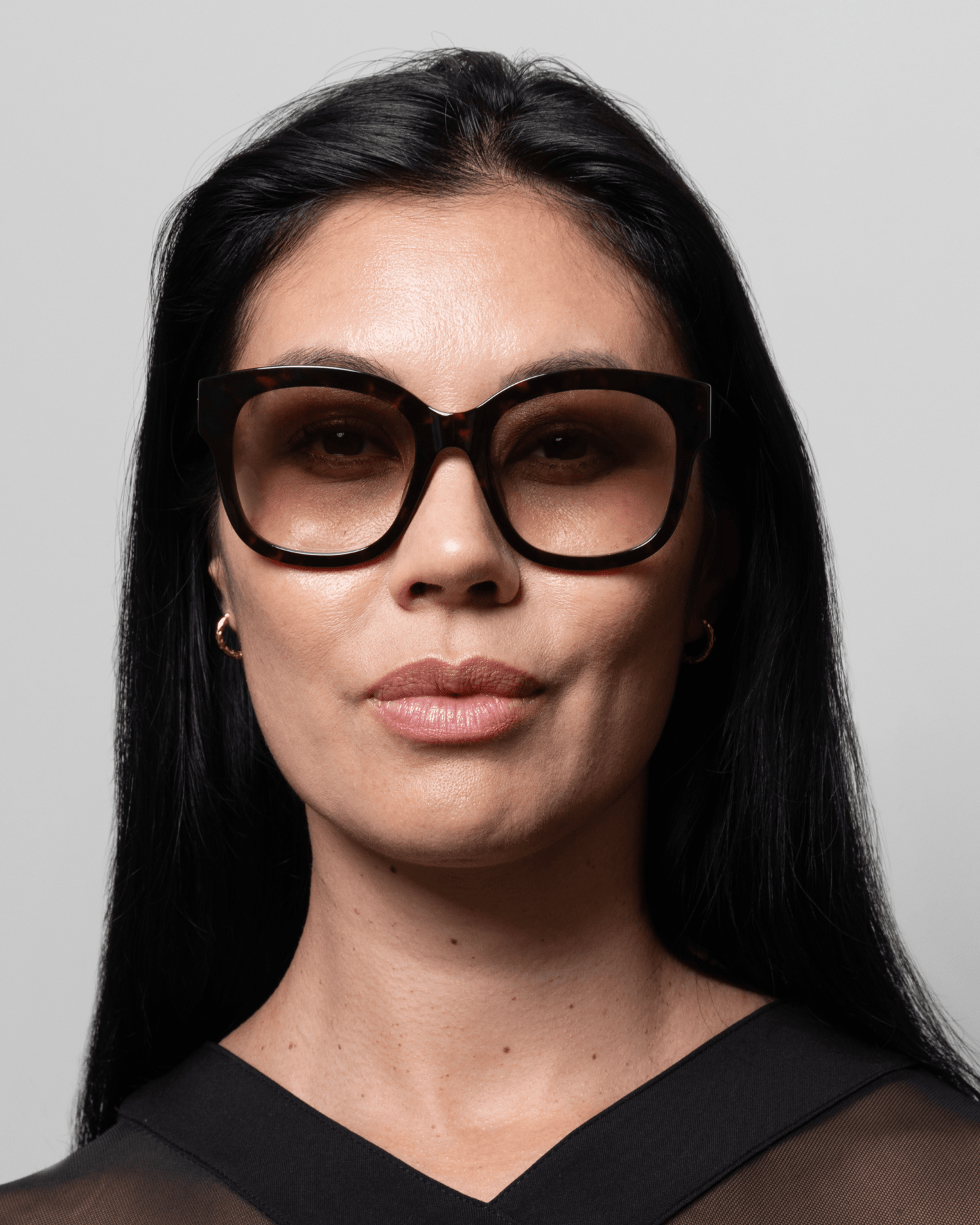 Kvinde med langt sort hår iført GLAS Kate Brun-mønstrede læsebriller, små hoop-øreringe, sort top og grå baggrund.
