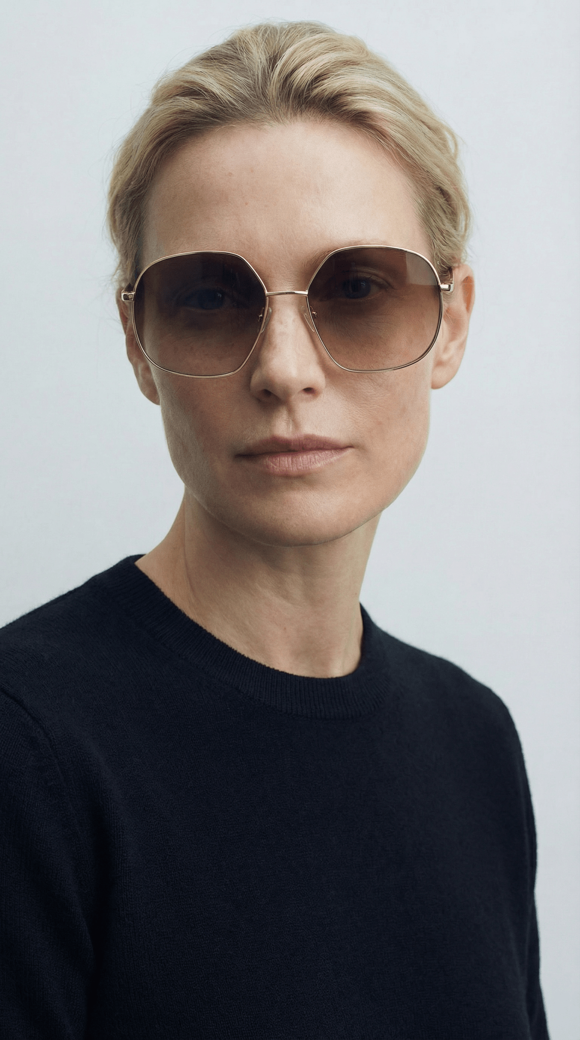 En person med kort, blondt hår har GLAS-solbrillerne »Cornelia Guld« på og en sort trøje med rund hals, og ser fremad.