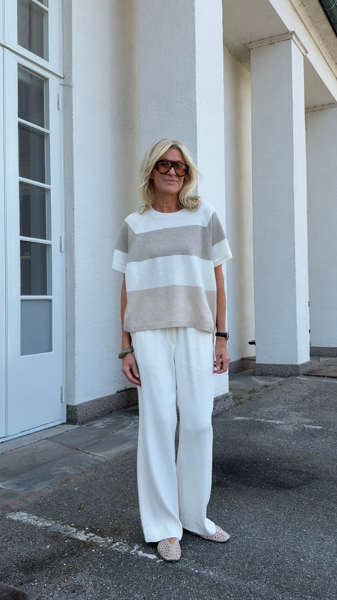 En kvinde står udenfor iført en »Gustav – Celma Off white strikbluse dame«, solbriller, hvide bukser og flade sko.
