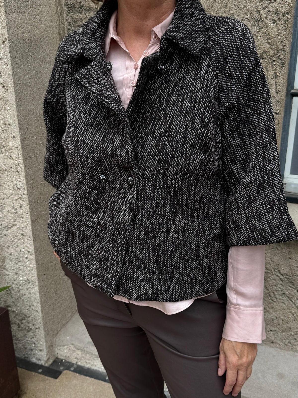 En person iført en Gustav – Nora Brunmeleret-jakke over en lyserød skjorte og brune bukser, der står ved en stenmur.