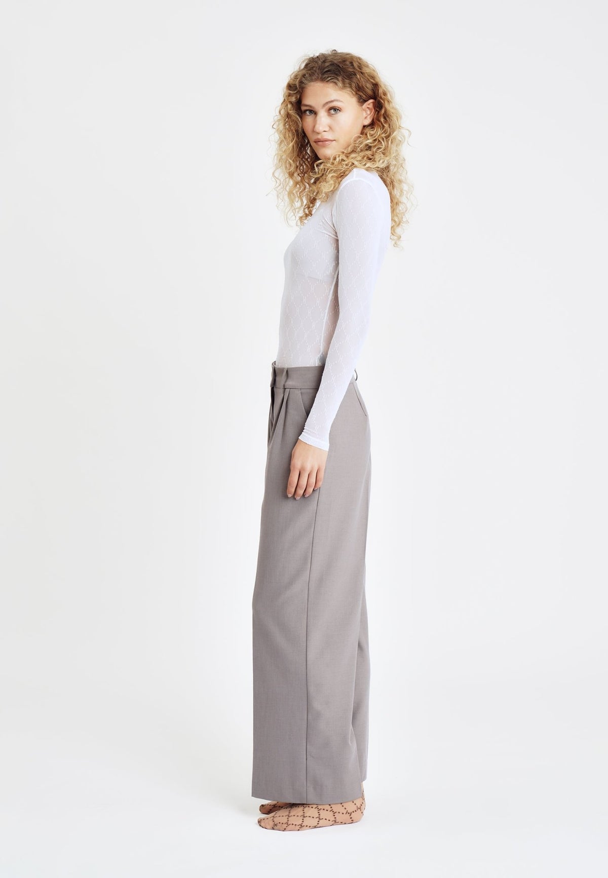 Kvinde, der står med siden til, iført en HYPE THE DETAIL-meshbluse i offwhite, grå bukser med brede ben og brune mønstrede sko.