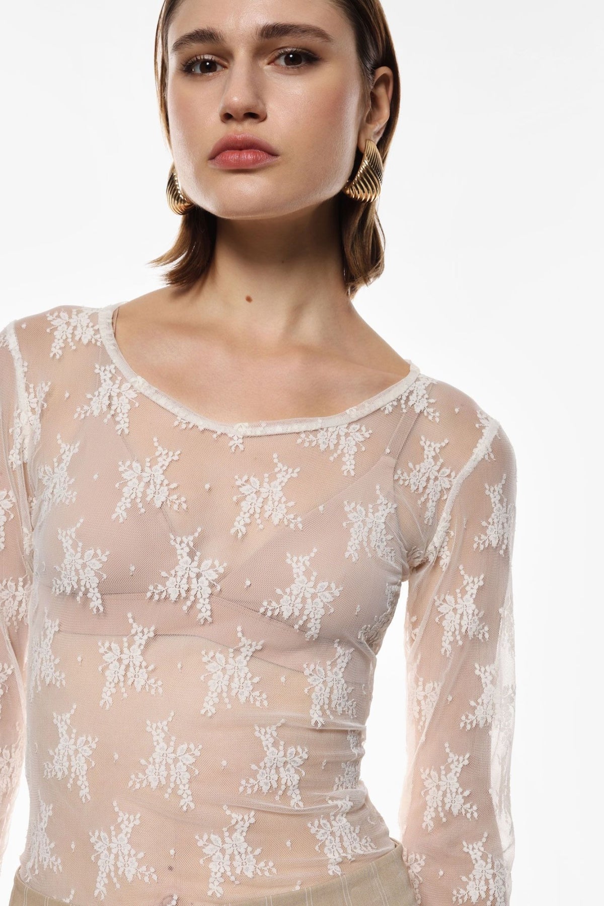 En kvinde iført en Imperial 152581 TW91 off-white, broderet, gennemsigtig langærmet bluse står foran en ensfarvet baggrund.