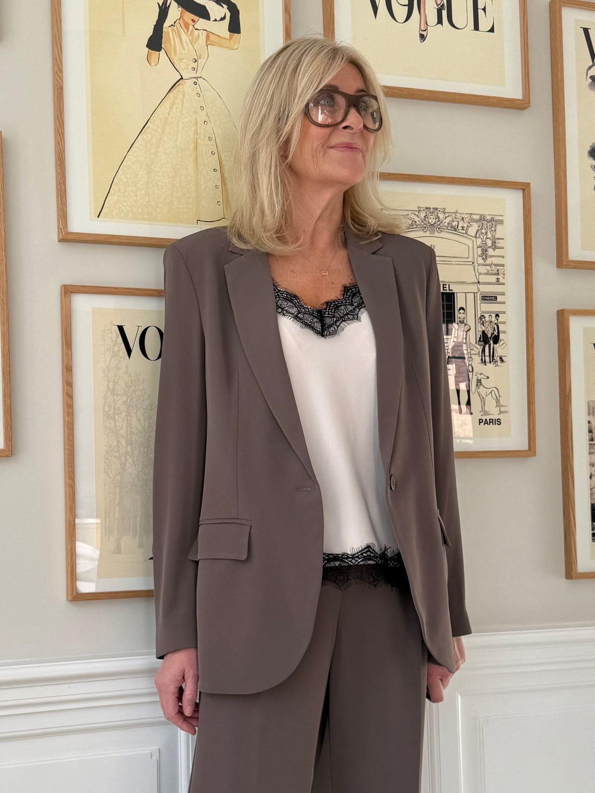 Kvinde i Imperial – JV96 gråbrun blazer og briller står ved siden af indrammede Vogue-forsider på væggen.