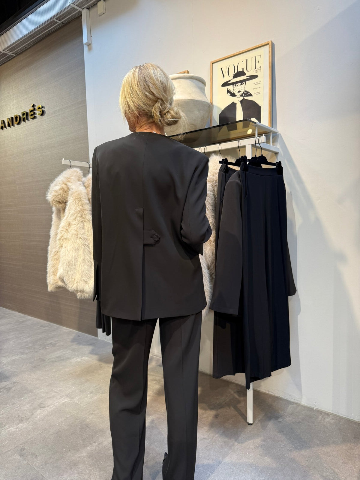 En blond person i Imperial – JCA9 Kaffe-blazer – kigger på tøj ved siden af en Vogue-plakat og en stor keramisk vase.