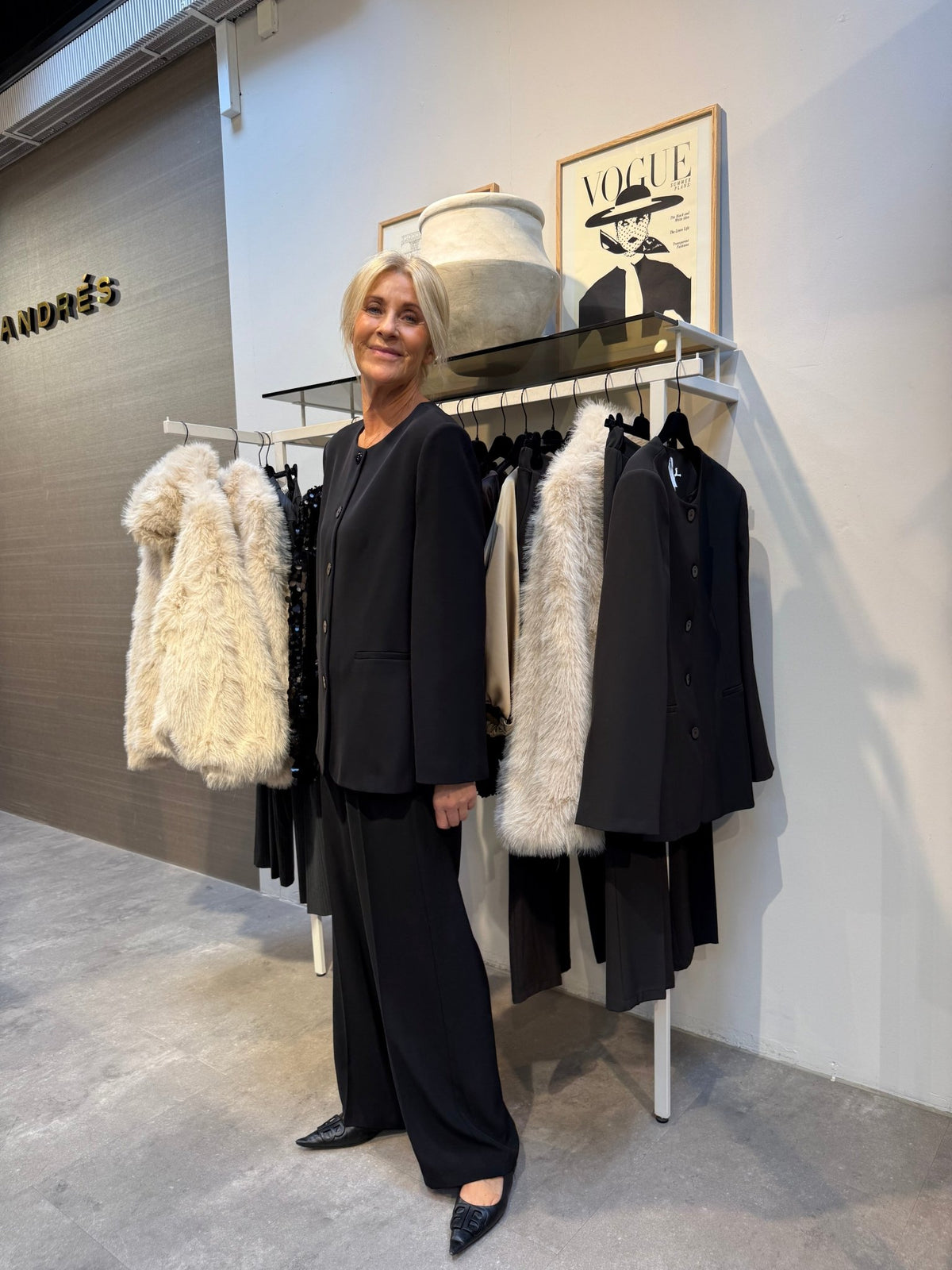 En kvinde iført en Imperial 155964 JCA9 Sort-blazer står i en tøjbutik ved siden af frakker, indrammede kunstværker og en vase, der står ovenover.
