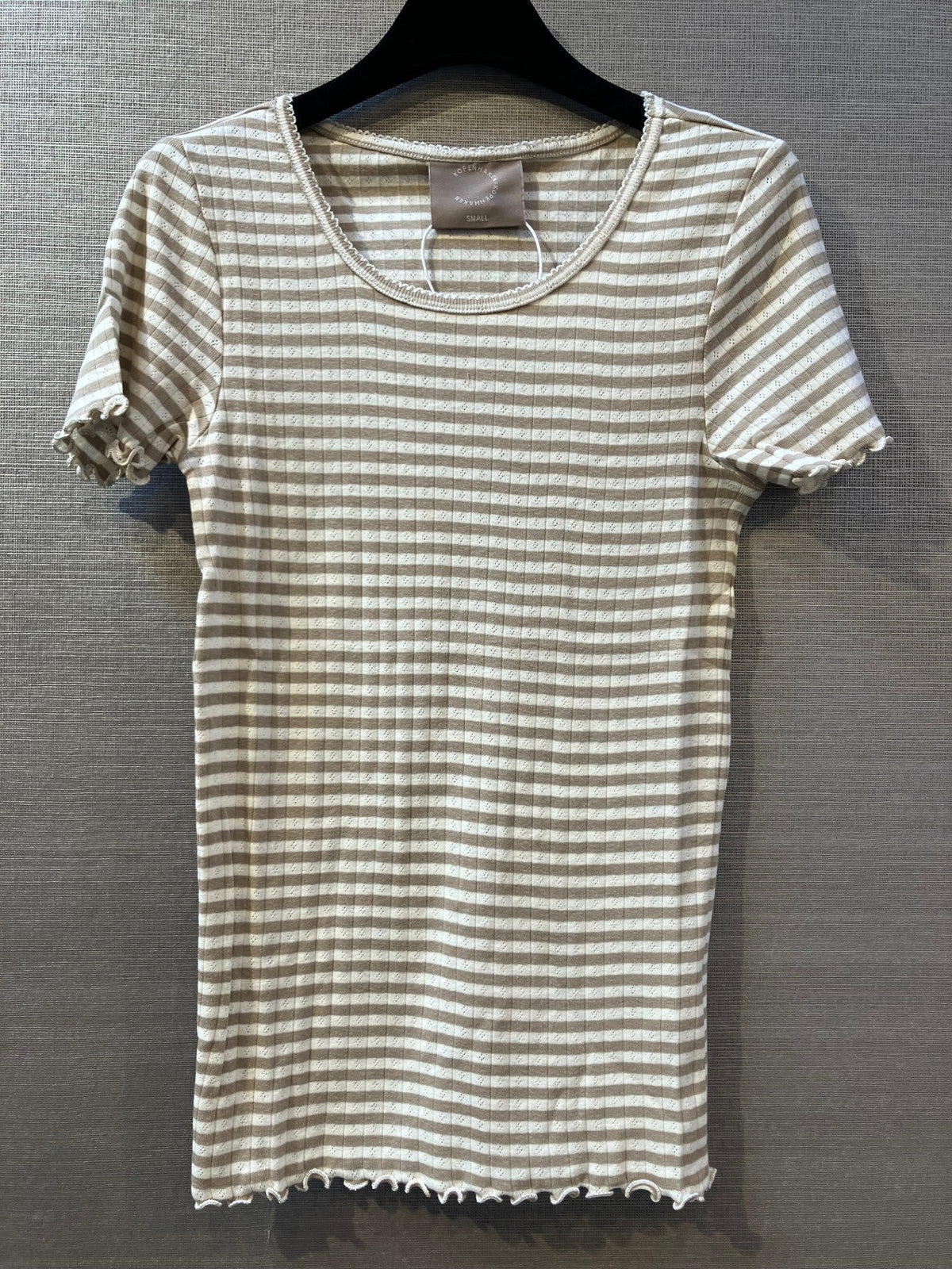 Kopenhaken 154480 Blød T-shirt i lysebrun med tan- og hvide striber på en sort bøjle foran en grå, struktureret væg.