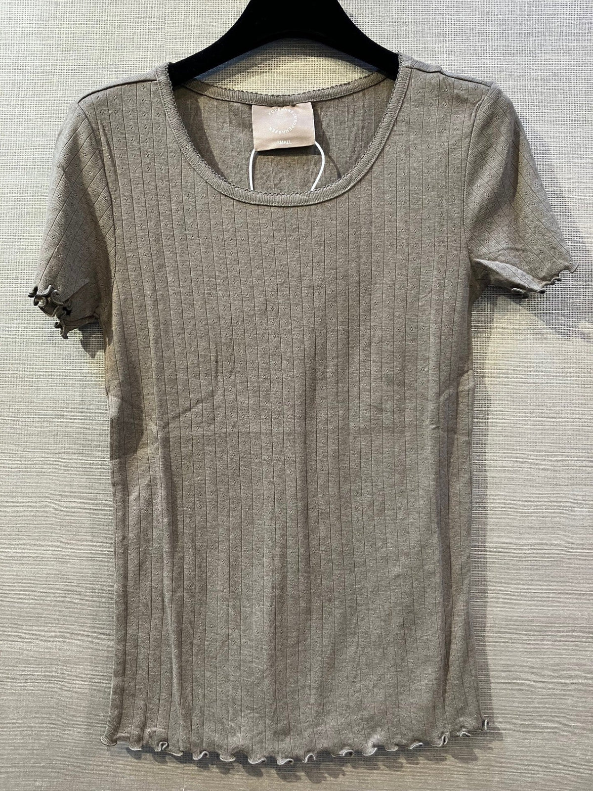 Kopenhaken Soft Tee Brun på bøjle; den ribstrikkede bomulds-T-shirt træder tydeligt frem mod den strukturerede væg.