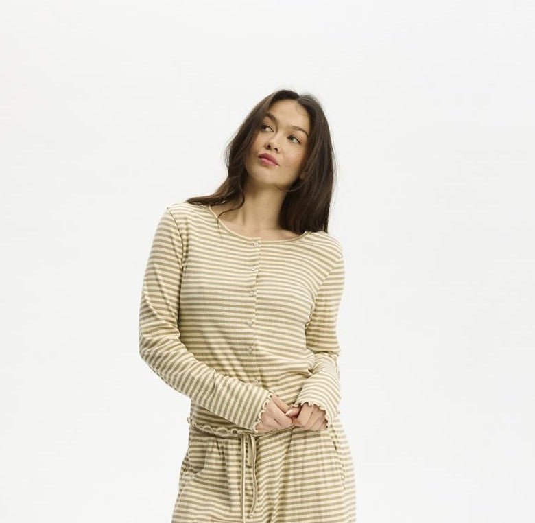 En kvinde er iført Kopenhakens »Soft Stripe Lysebrun«-sæt og poserer i beige og hvidt foran en ensfarvet hvid baggrund.