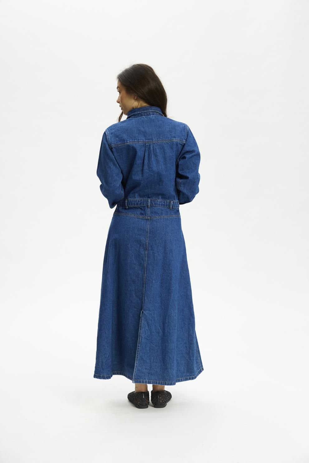 En person, der står med ryggen til kameraet, iført en Kopenhaken Osaka Denim-kjole og sorte sko, foran en ensfarvet hvid baggrund.