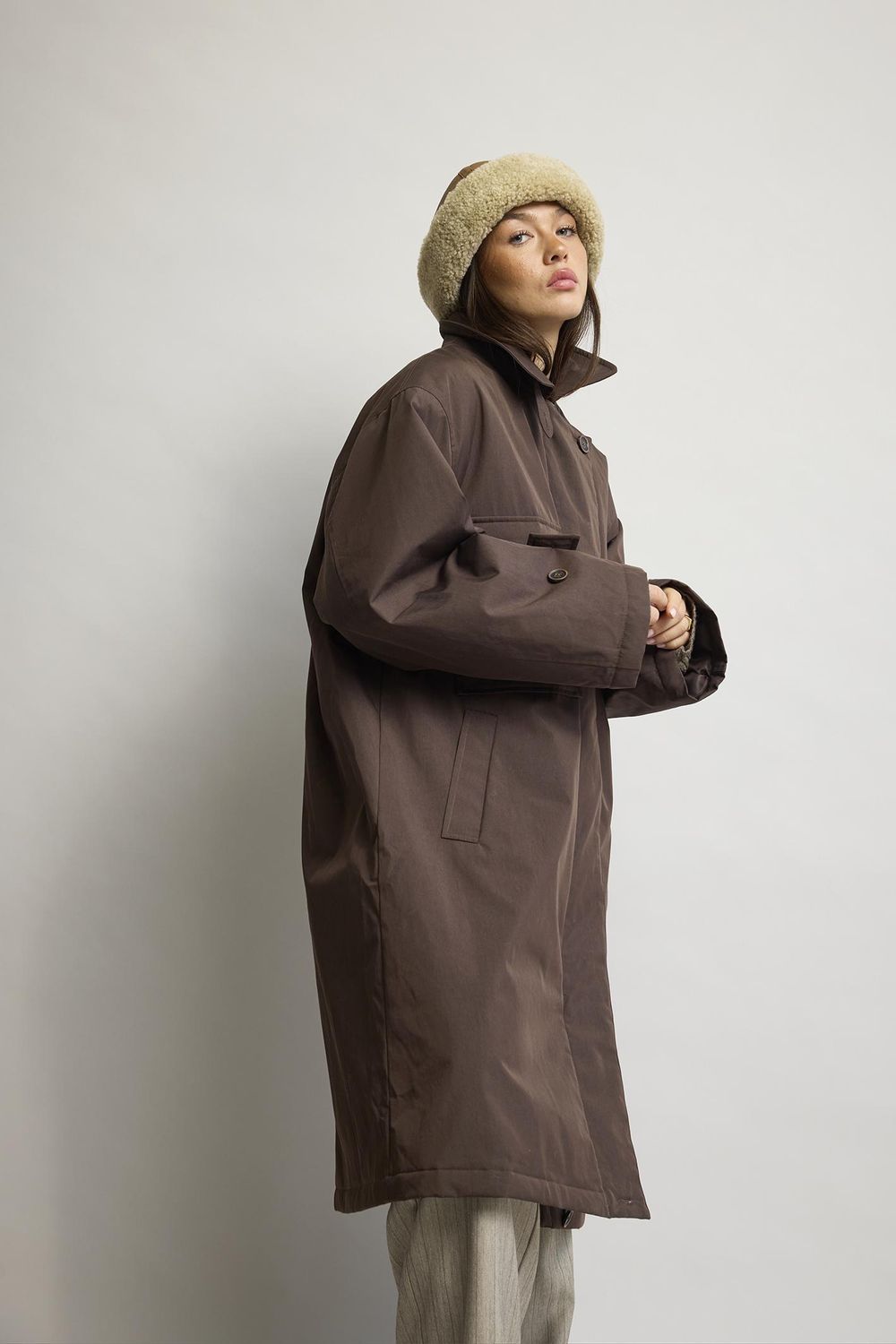 En person iført en Manchester Kaffe-overshirt fra Kopenhaken, en beige, lodden hue og grå bukser står foran en ensfarvet baggrund.