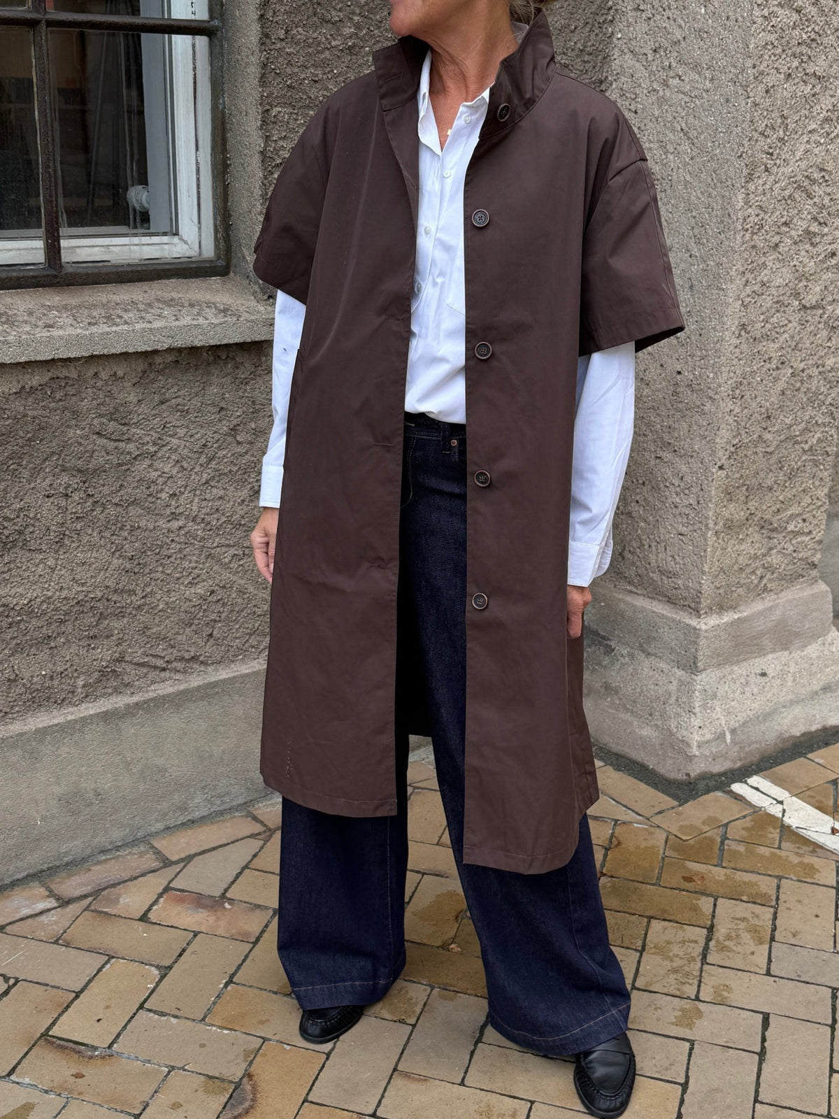 En person i hvid skjorte, brede mørke jeans og Kopenhaken MANCHESTER Kaffe står på en fortov af mursten ved et vindue.