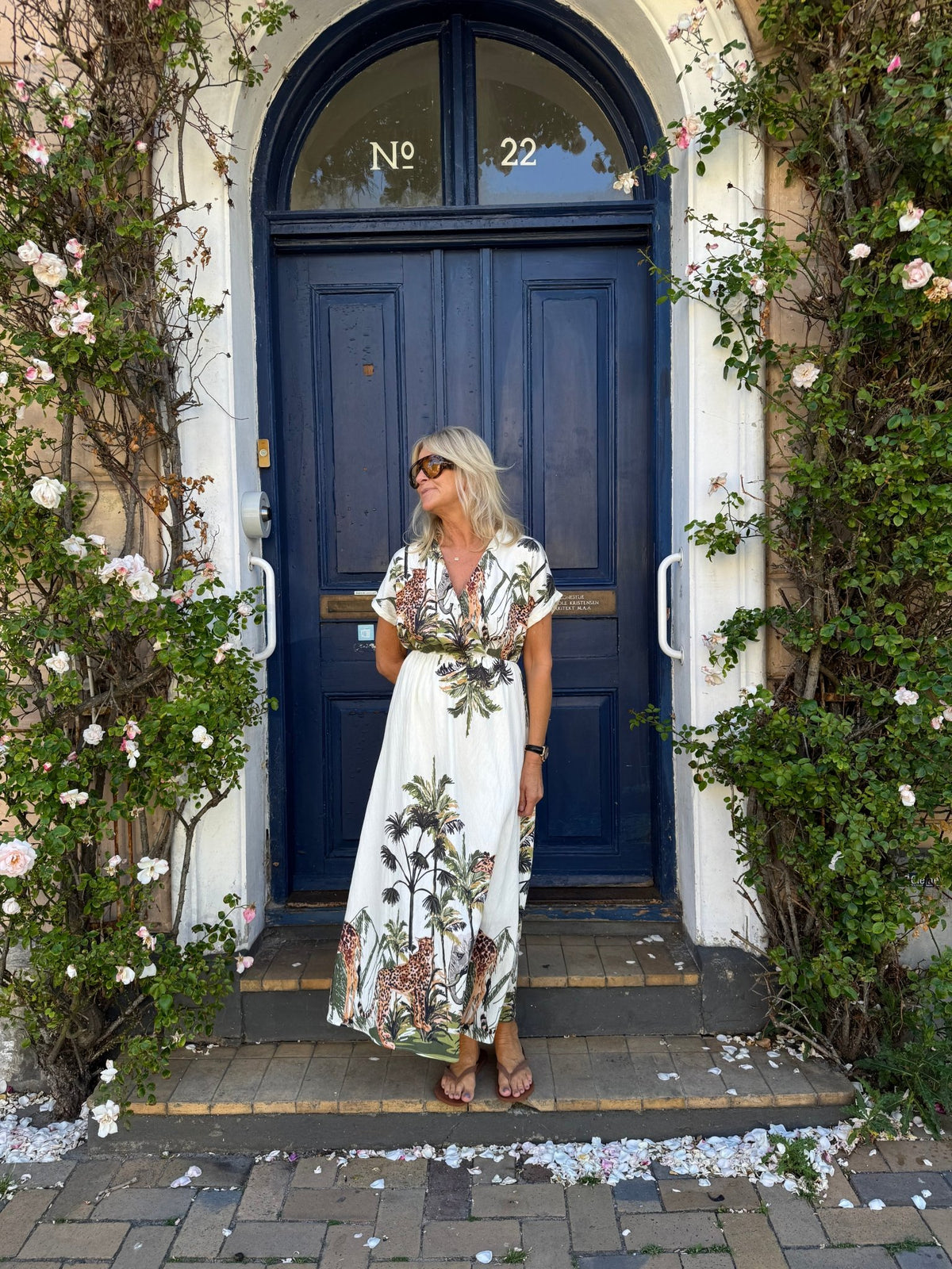 En kvinde i en mønstret kjole fra Luxzuz Via Off-White står foran en blå dør, omgivet af grønne ranker og hvide blomster.