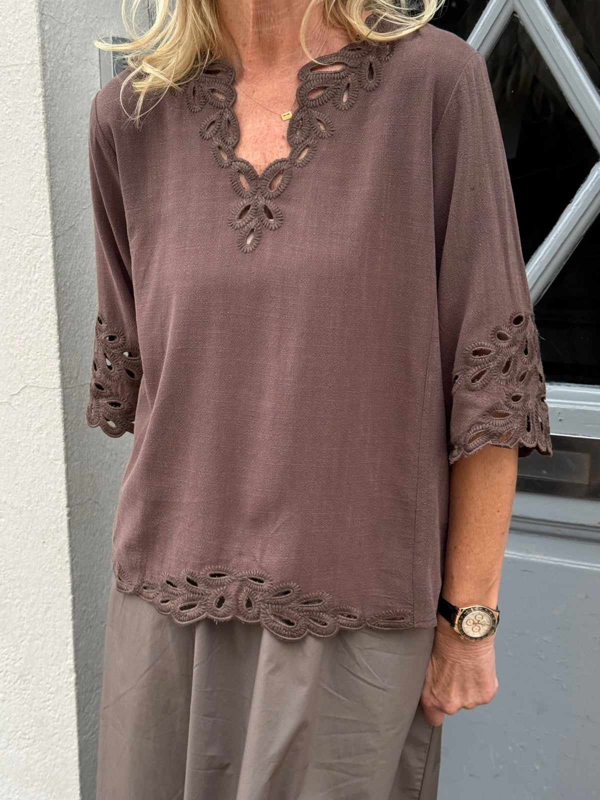 En person har Luxzuz Vicha Brun på, en mørkebrun bluse med korte ærmer og broderie anglaise-kant hele vejen rundt.