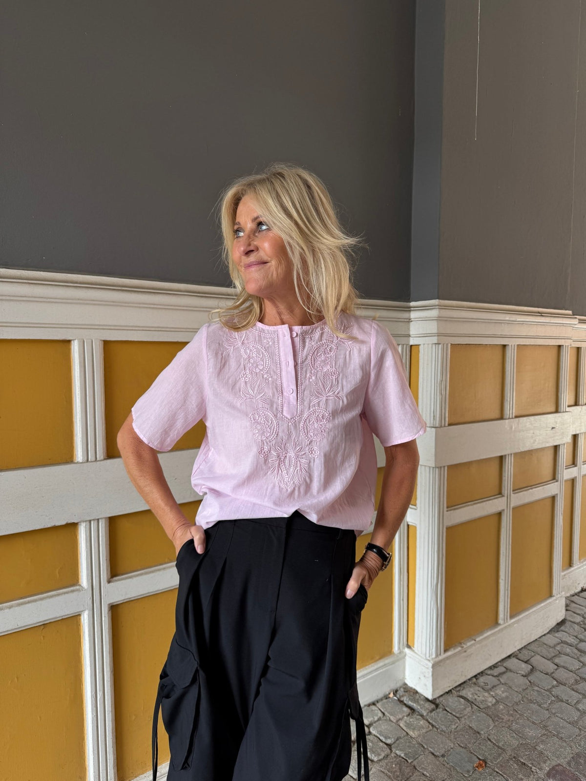 En blond kvinde i en Luxzuz – Viva Pink-bluse står indendørs ved en væg med gule og hvide paneler.