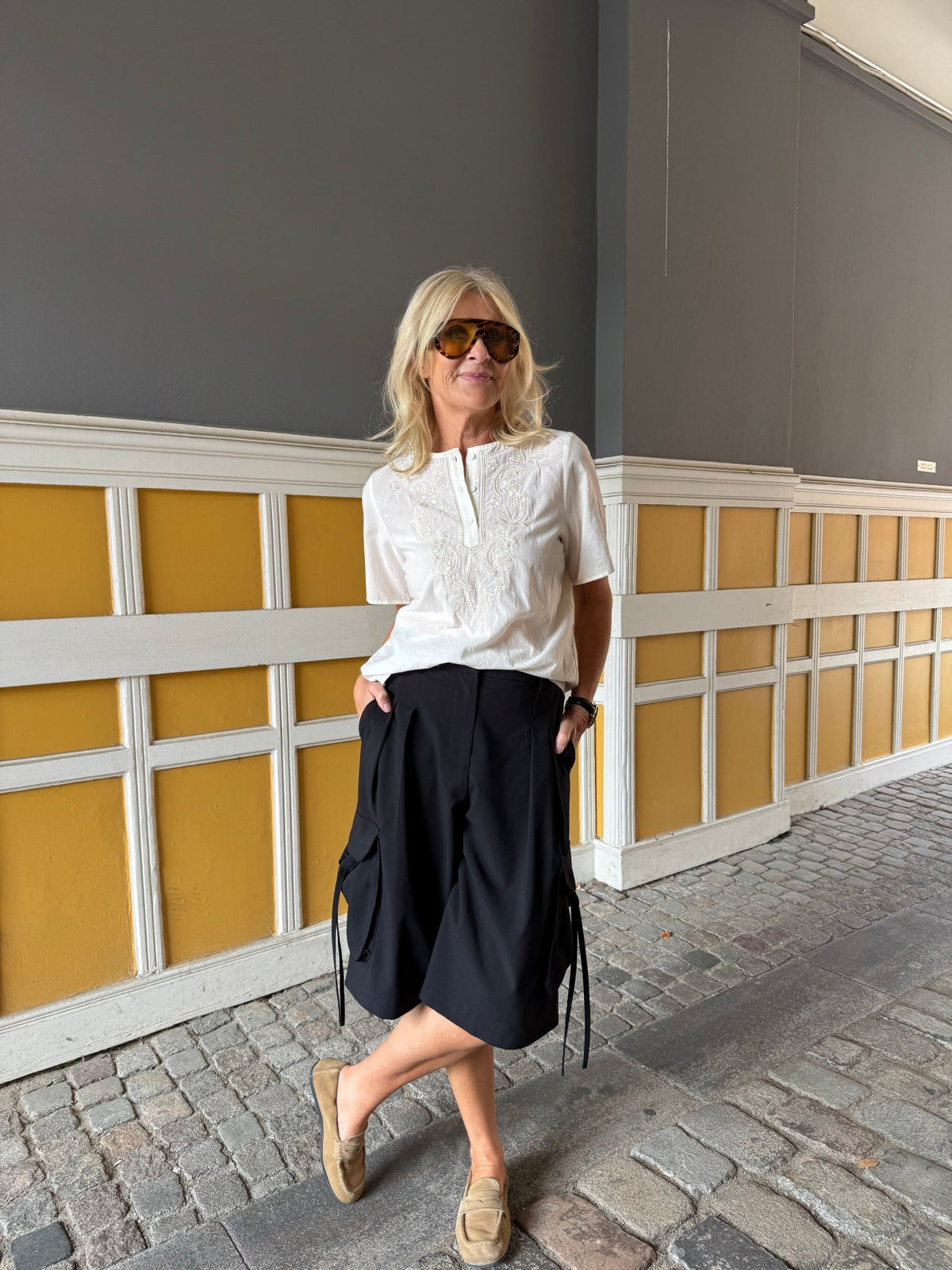 En kvinde med solbriller, en Luxzuz Viva Off White-bluse, et sort knælængt nederdel og loafers står ved en gul væg.