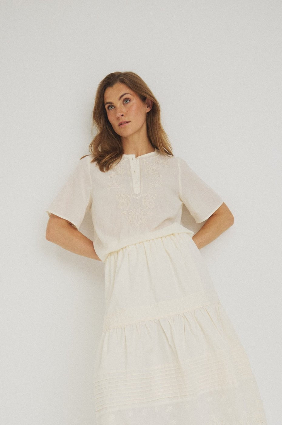 En kvinde iført en Luxzuz Viva-bluse i offwhite og et lagdelt nederdel, der står foran en ensfarvet hvid baggrund.