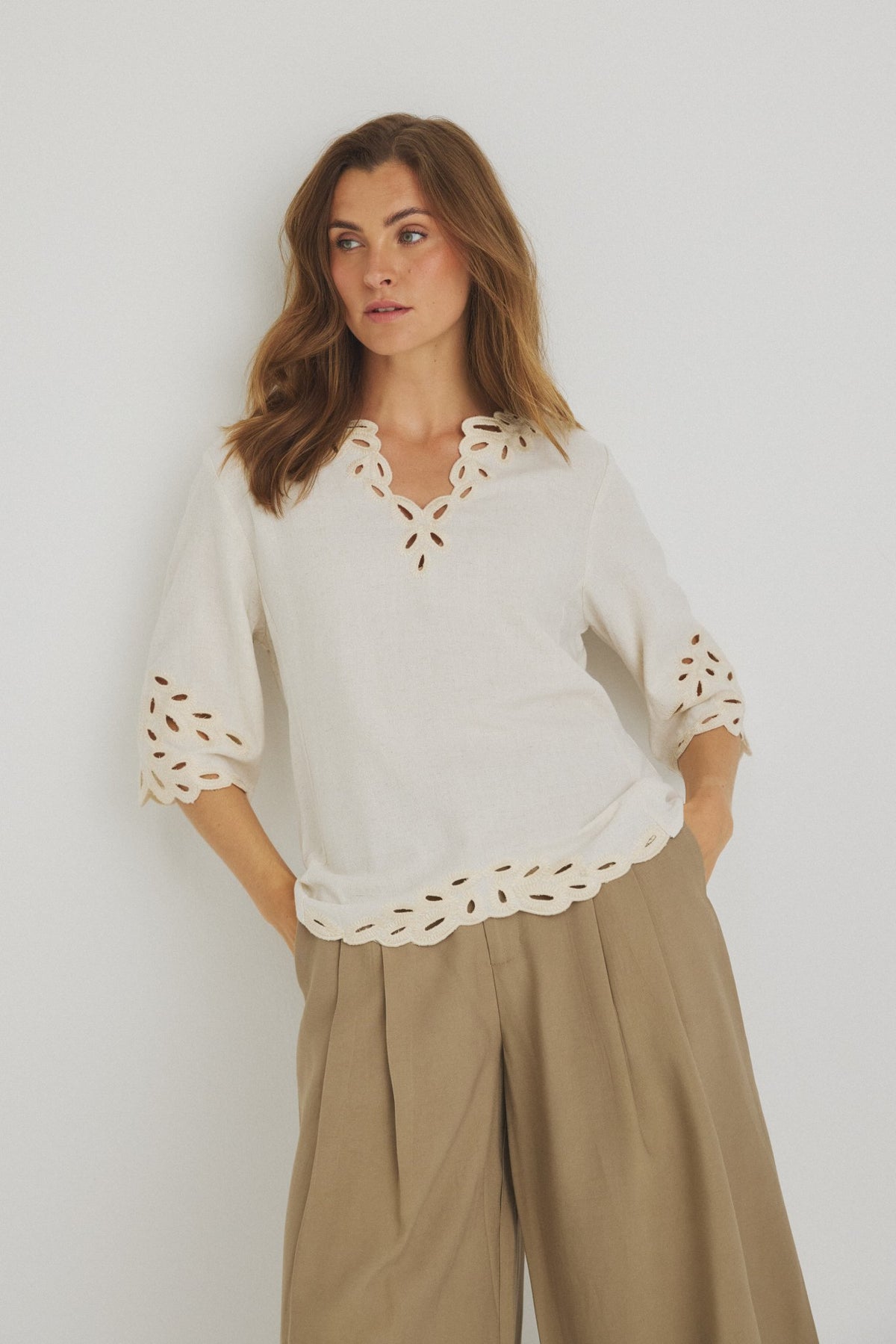 En kvinde er iført en offwhite Luxzuz Vicha-bluse og beige bukser med brede ben og står foran en ensfarvet hvid baggrund.