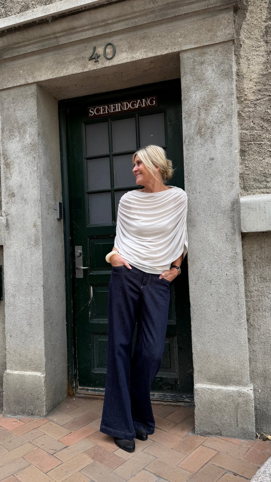 En kvinde iført en offwhite Luxzuz Queenie-top og mørke bukser står afslappet ved en grøn dør med skiltet »SCENEINGANG«.