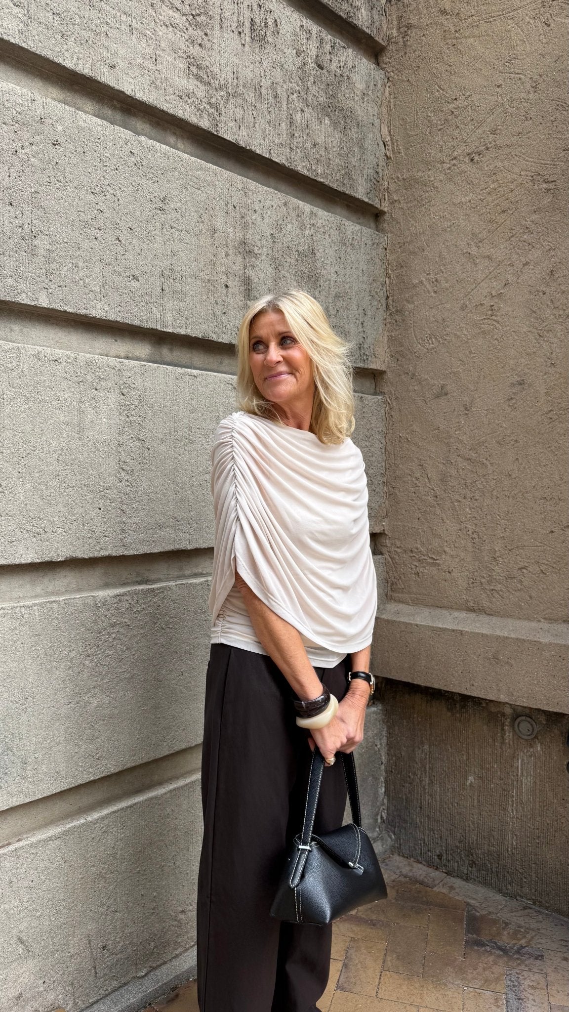 En blond kvinde iført en Luxzuz Queenie Off White-bluse, mørke bukser og en sort håndtaske står ved en betonmur.