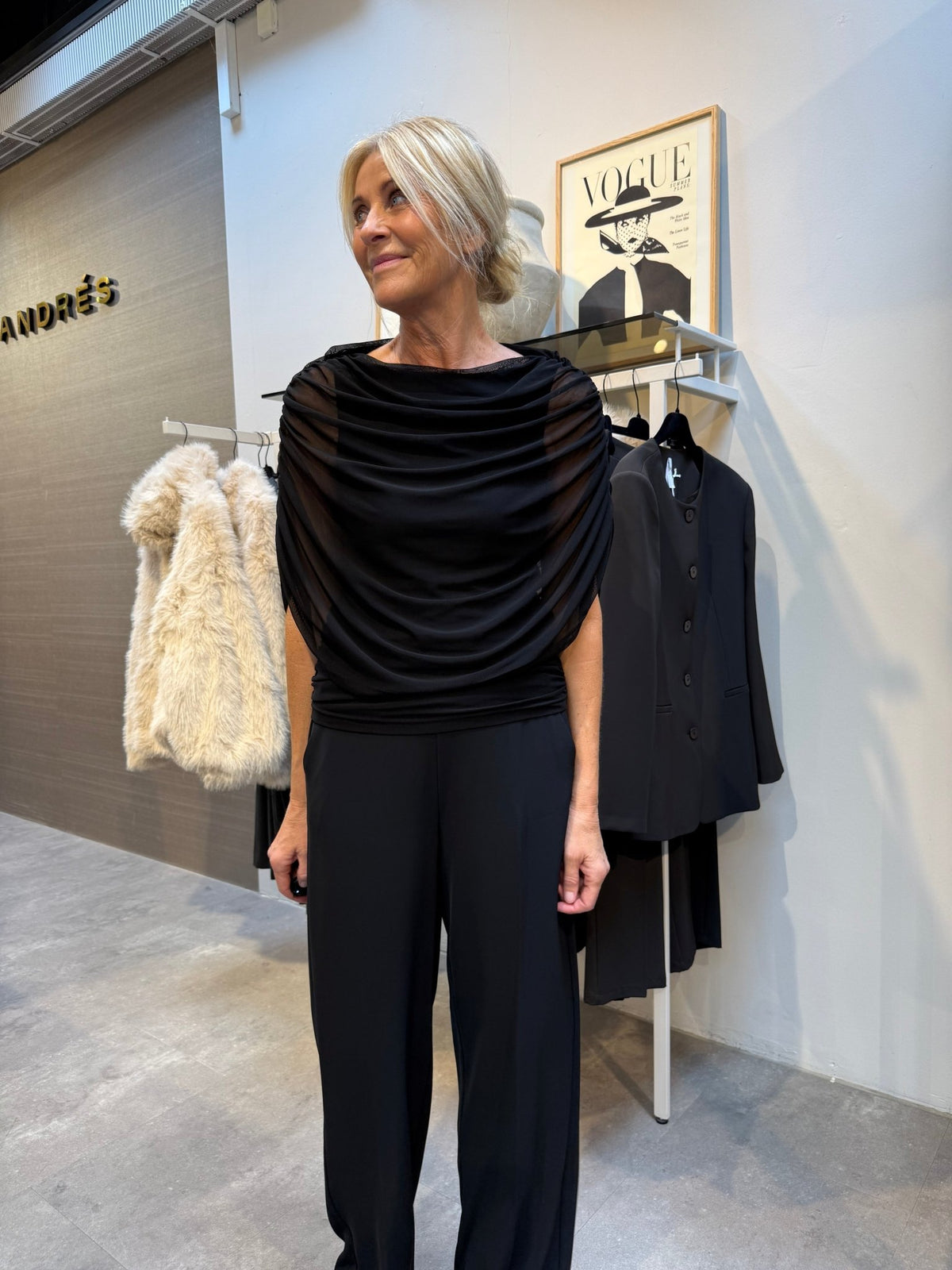 En kvinde i sort har en Luxzuz Queenie Sort-bluse på og står ved siden af nogle jakker, en pelsfrakke og et indrammet Vogue-forsidebillede.
