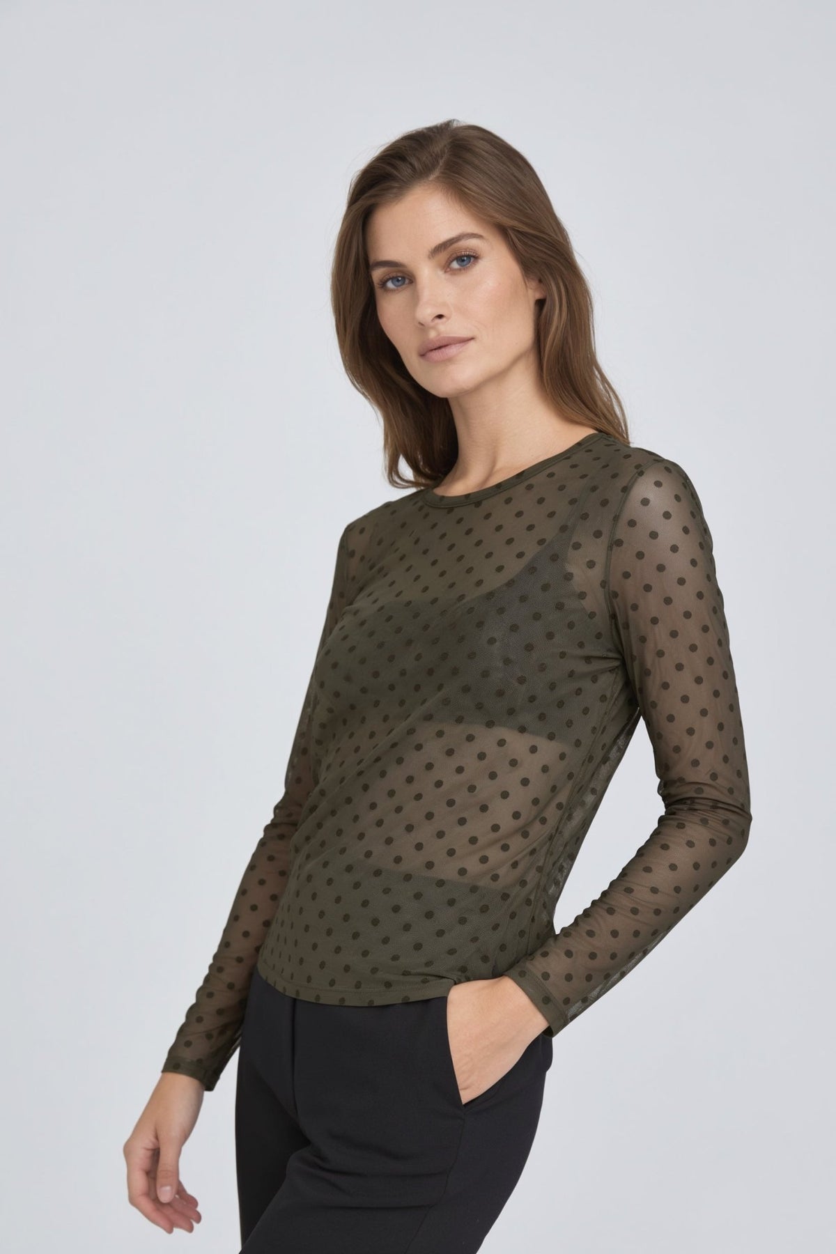 En kvinde har en Luxzuz 156326 - Dot Mesh Army grøn, gennemsigtig, langærmet olivengrøn bluse med mørke prikker på sig.