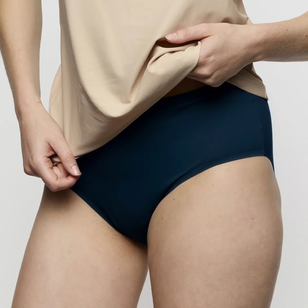 En person i en beige top løfter trøjen og afslører Marie Jo – Soft Studio Mørkeblå 155155, et maxi-undertøj med høj talje.
