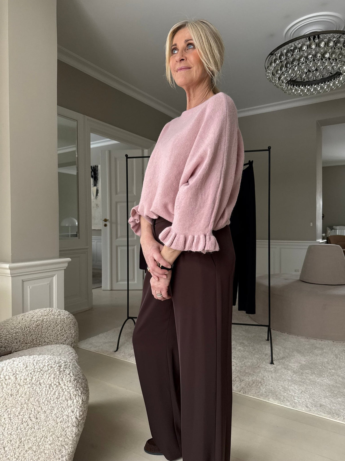 En kvindelig model viser Marta du Château Elderflower Knit Gammelrosa i lyserødt med brune bukser foran en neutral indendørs baggrund.