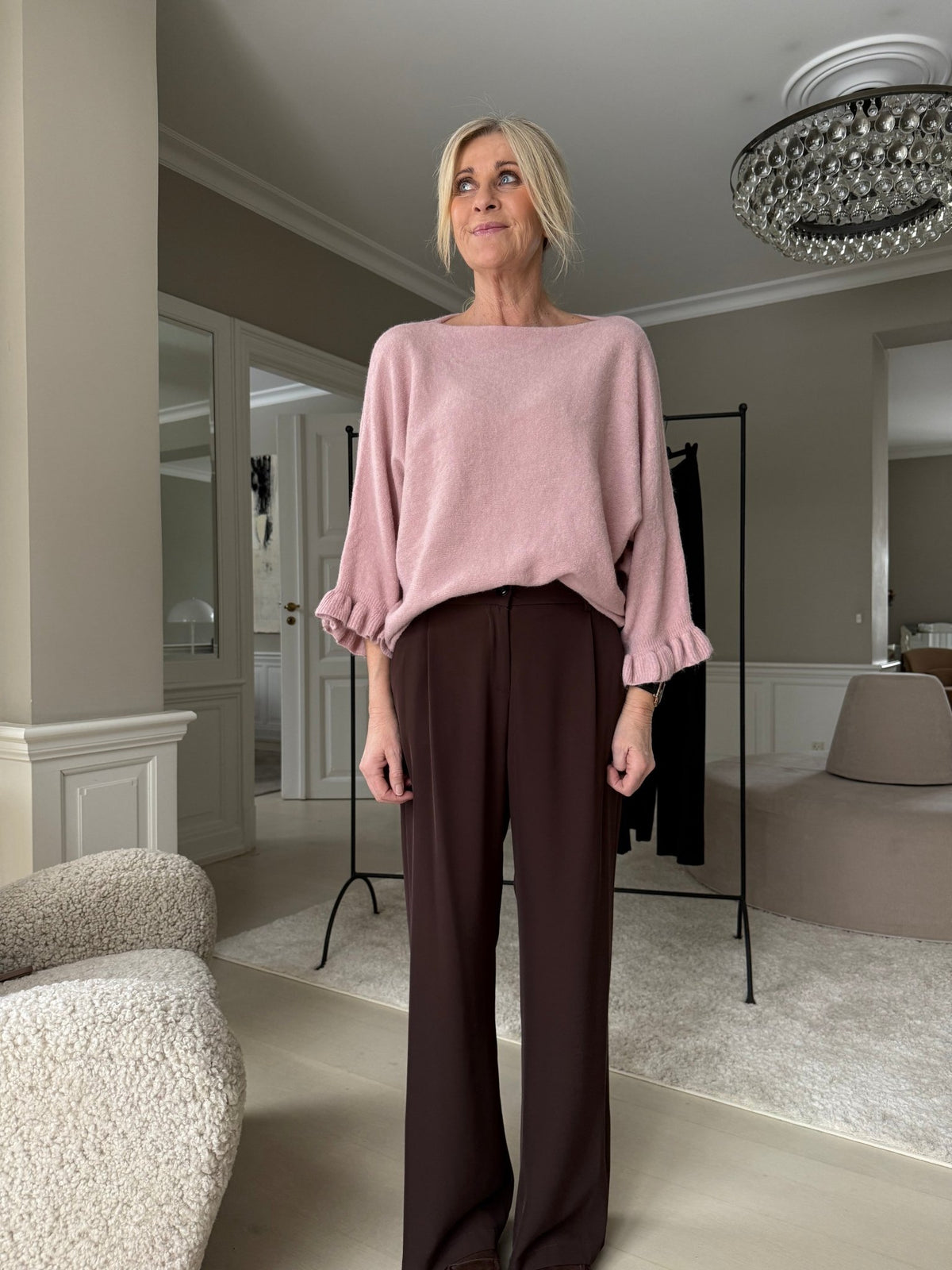 Kvinde indendørs iført Marta du Château-strik i farven »Elderflower Knit Gammelrosa«, brune bukser, moderne neutral baggrund.
