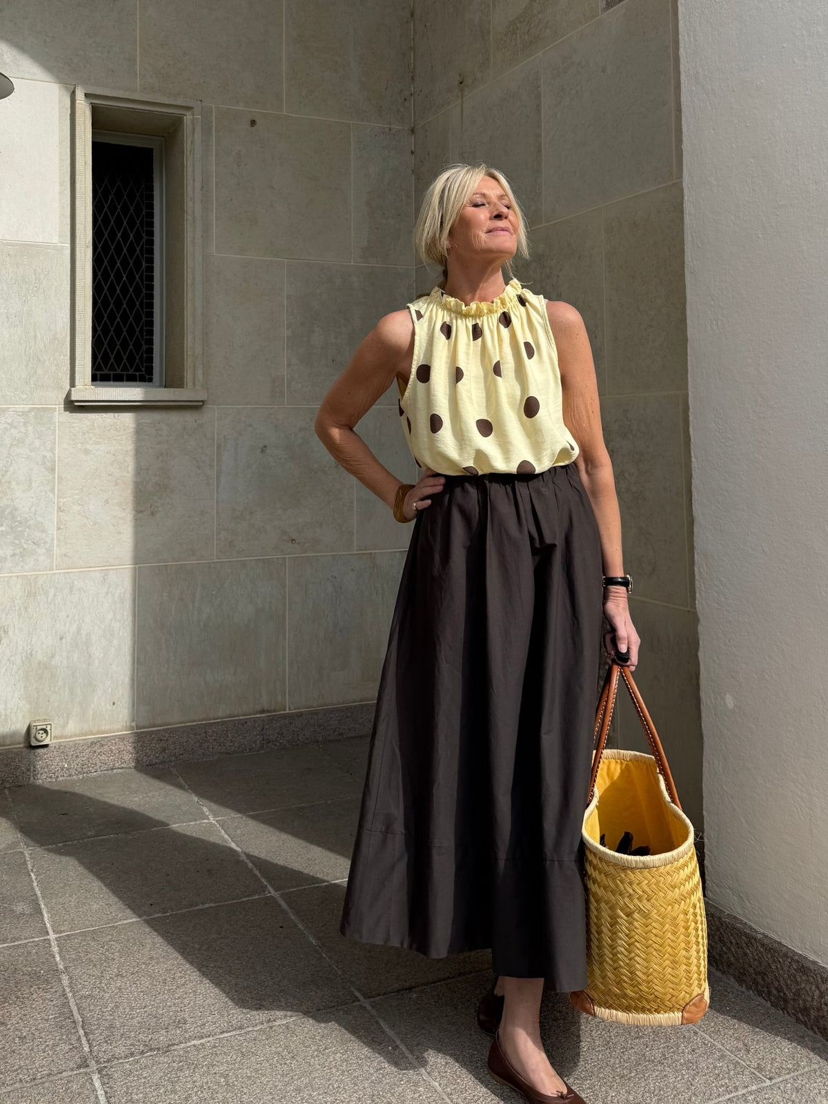 En kvinde i en top fra Marta du Château Lucina Gul, sort nederdel og en flettet taske står ved en solbeskinnet stenvæg.
