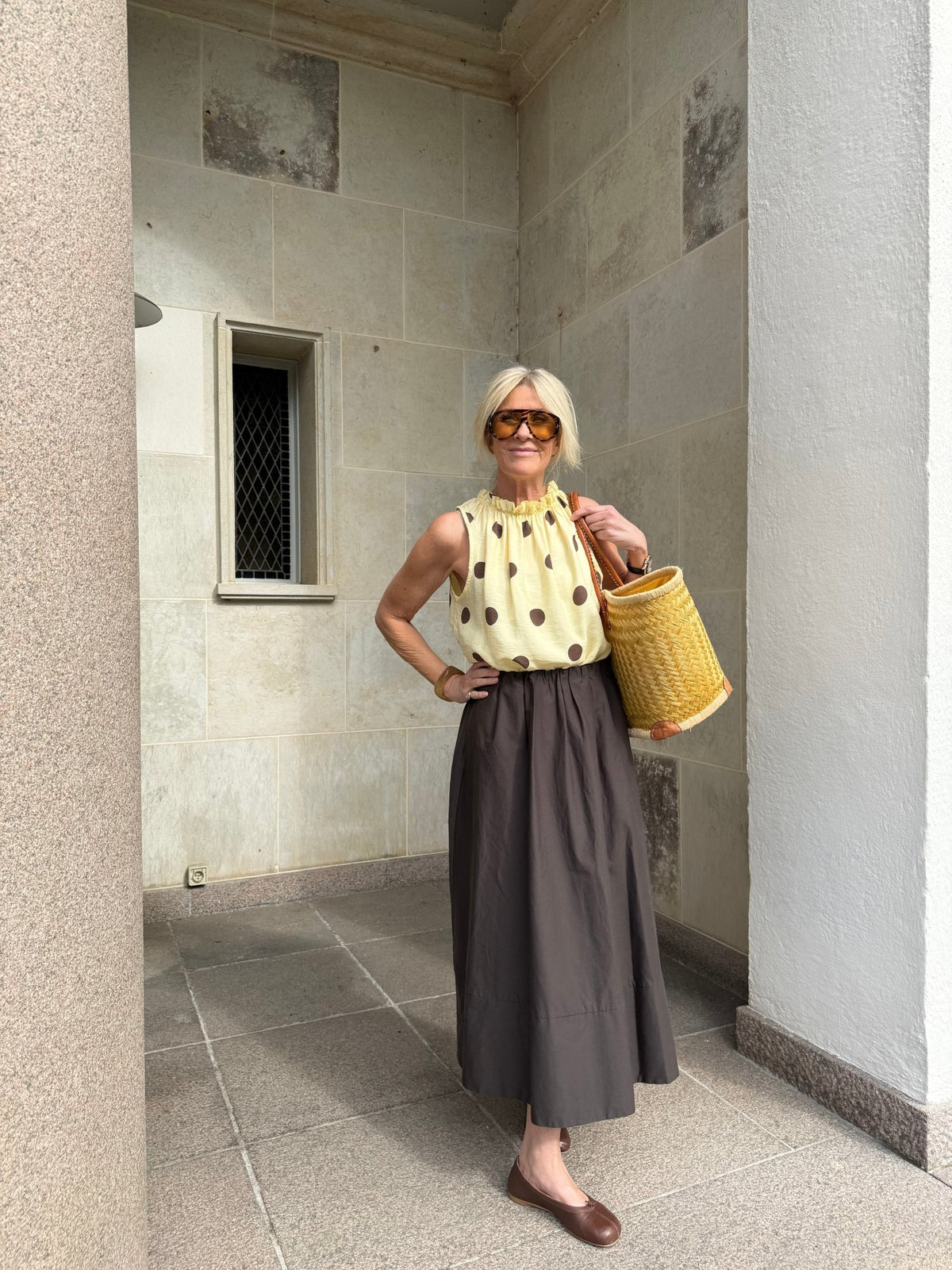 Kvinden med solbriller har en Marta du Château Lucina-top og en lang brun nederdel på og bærer en gul flettet Marta du Château-taske.