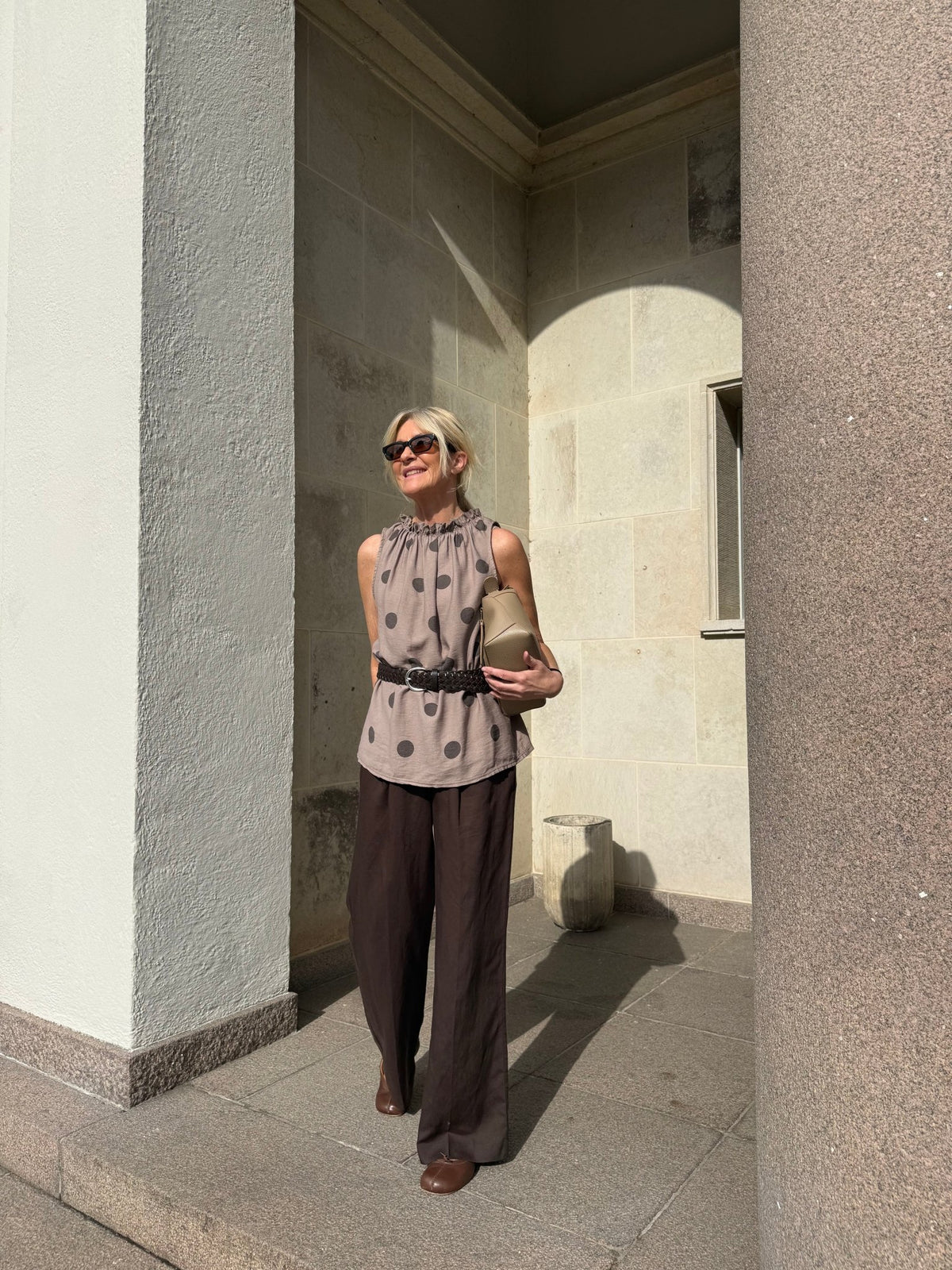 En kvinde med solbriller har en top fra Marta du Château Lucina Brun og brune bukser på og holder en beige clutch i hånden ved siden af en søjle.