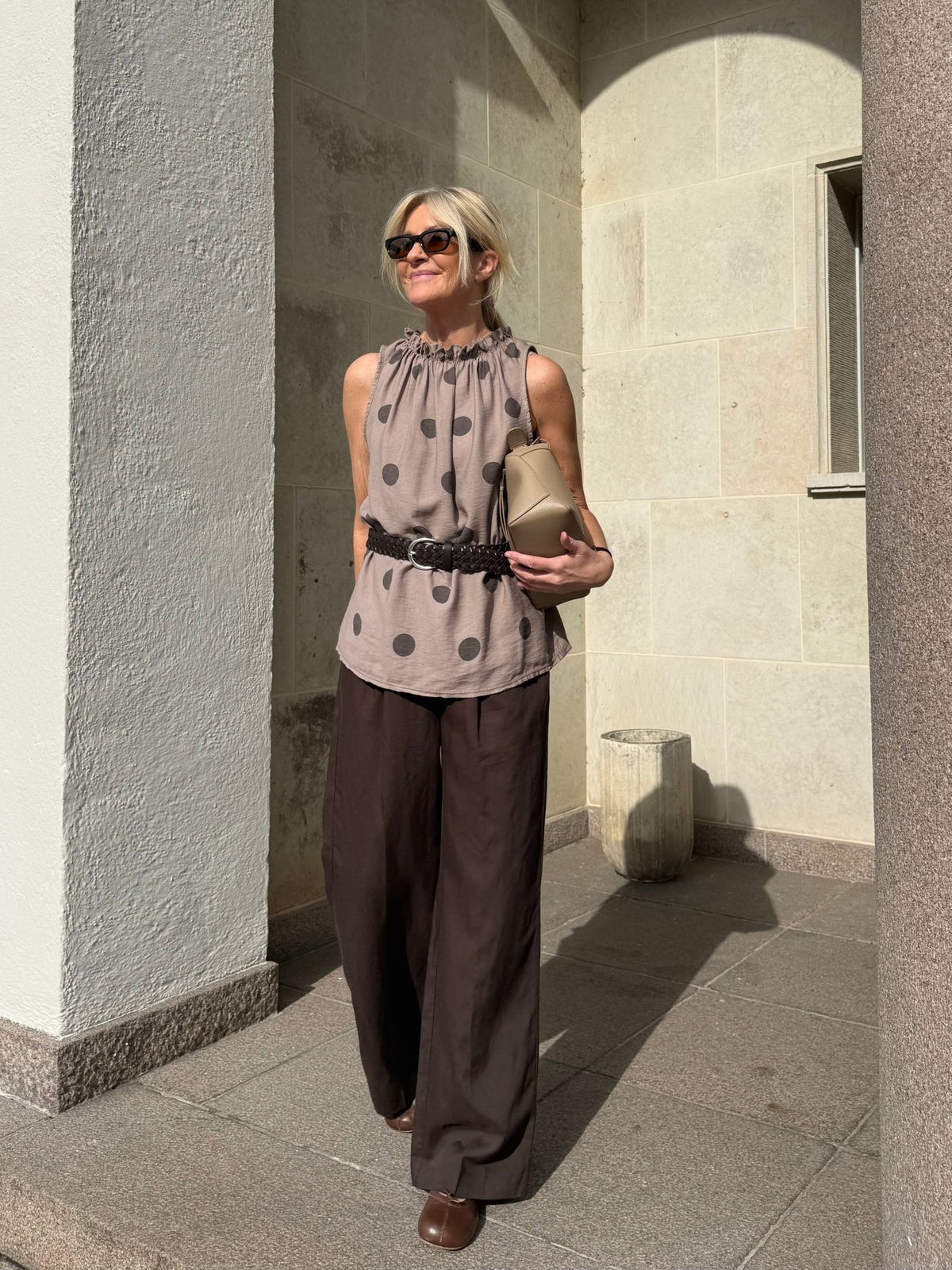 En kvinde med solbriller har en top fra Marta du Château Lucina Brun og brede brune bukser på og står udenfor med en beige clutch i hånden.
