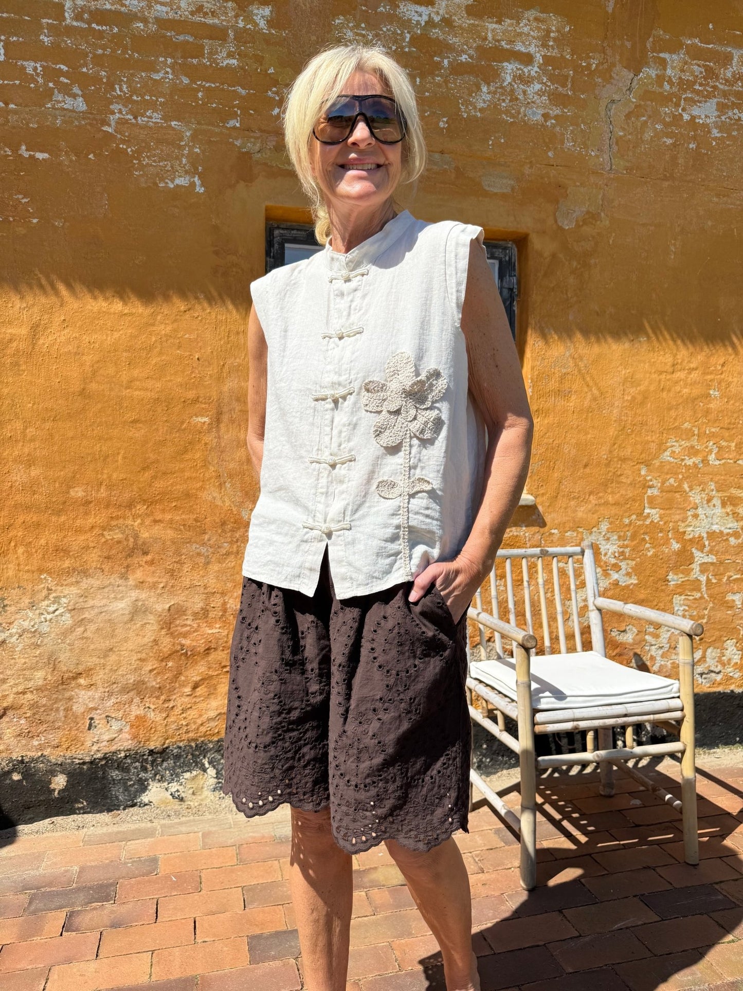 En kvinde iført en beige top fra Marta du Château Olina, solbriller og mørke shorts står ved en gul væg med struktur.