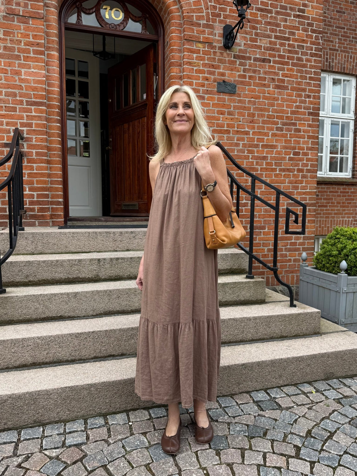 En kvinde i en løs kjole står på en stentrappe ved en murstensbygning og holder en Marta du Château Valisse Brun-håndtaske.
