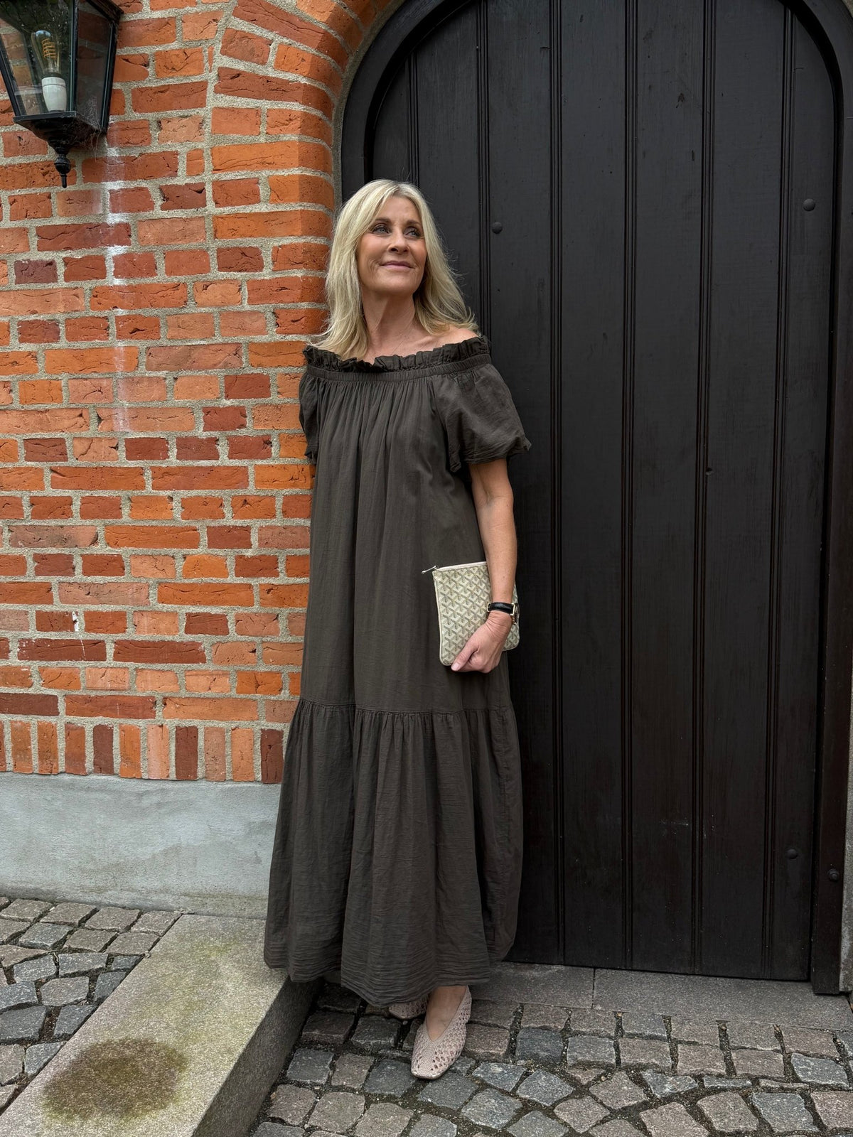 En kvinde i en mørkebrun Zenith-kjole fra Marta du Château står ved en sort dør på brosten og holder en beige clutch i hånden.