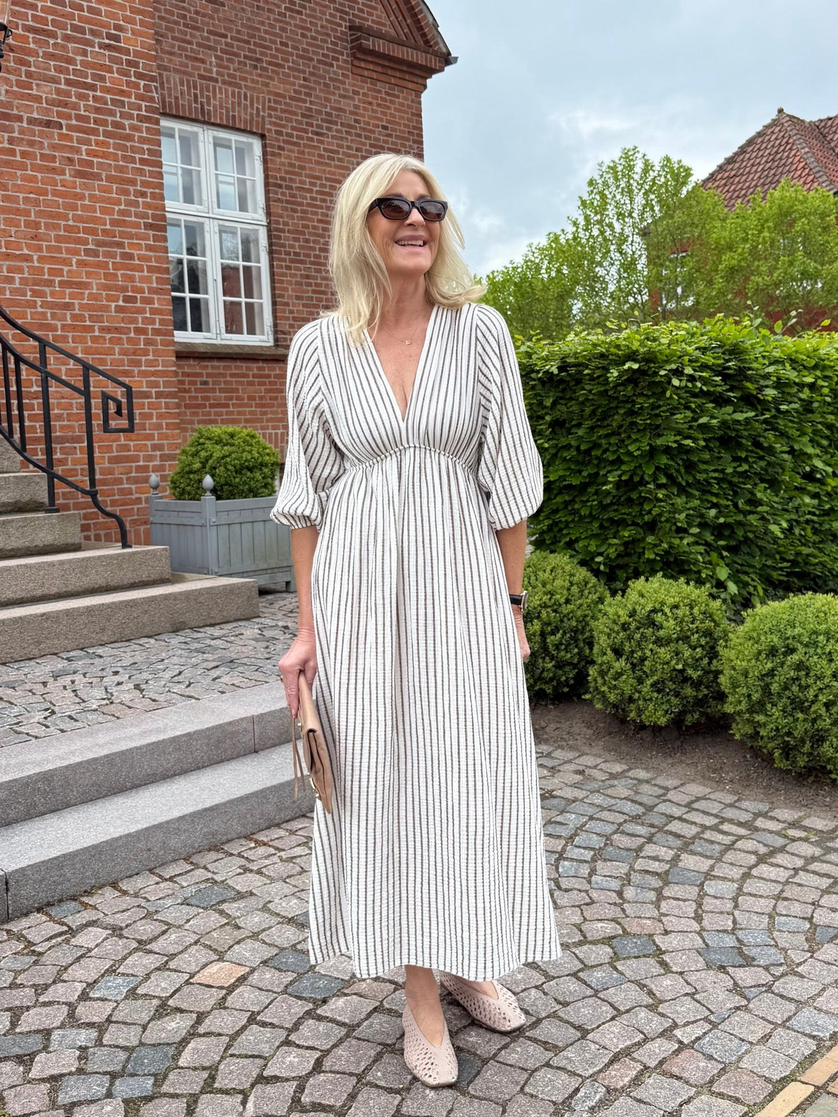 En kvinde i en stribet kjole fra Marta du Château Clario står på brosten, med en clutch i hånden og solbriller på.