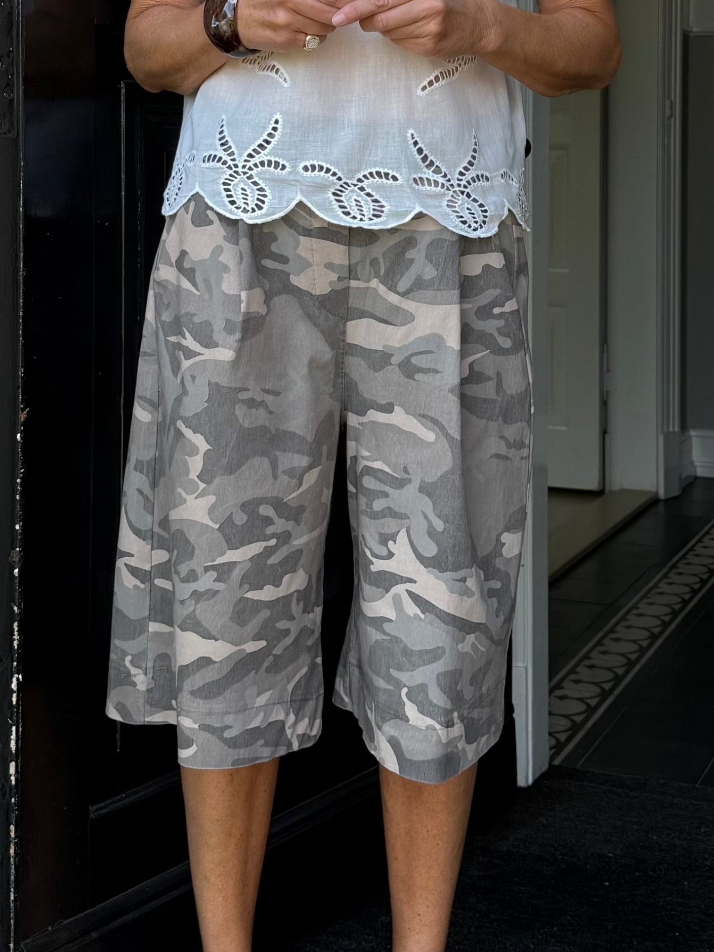 En person i en hvid bluse og grønne shorts fra Marta du Château Zuri Army står inde ved en døråbning, hvor hænderne er delvist synlige.