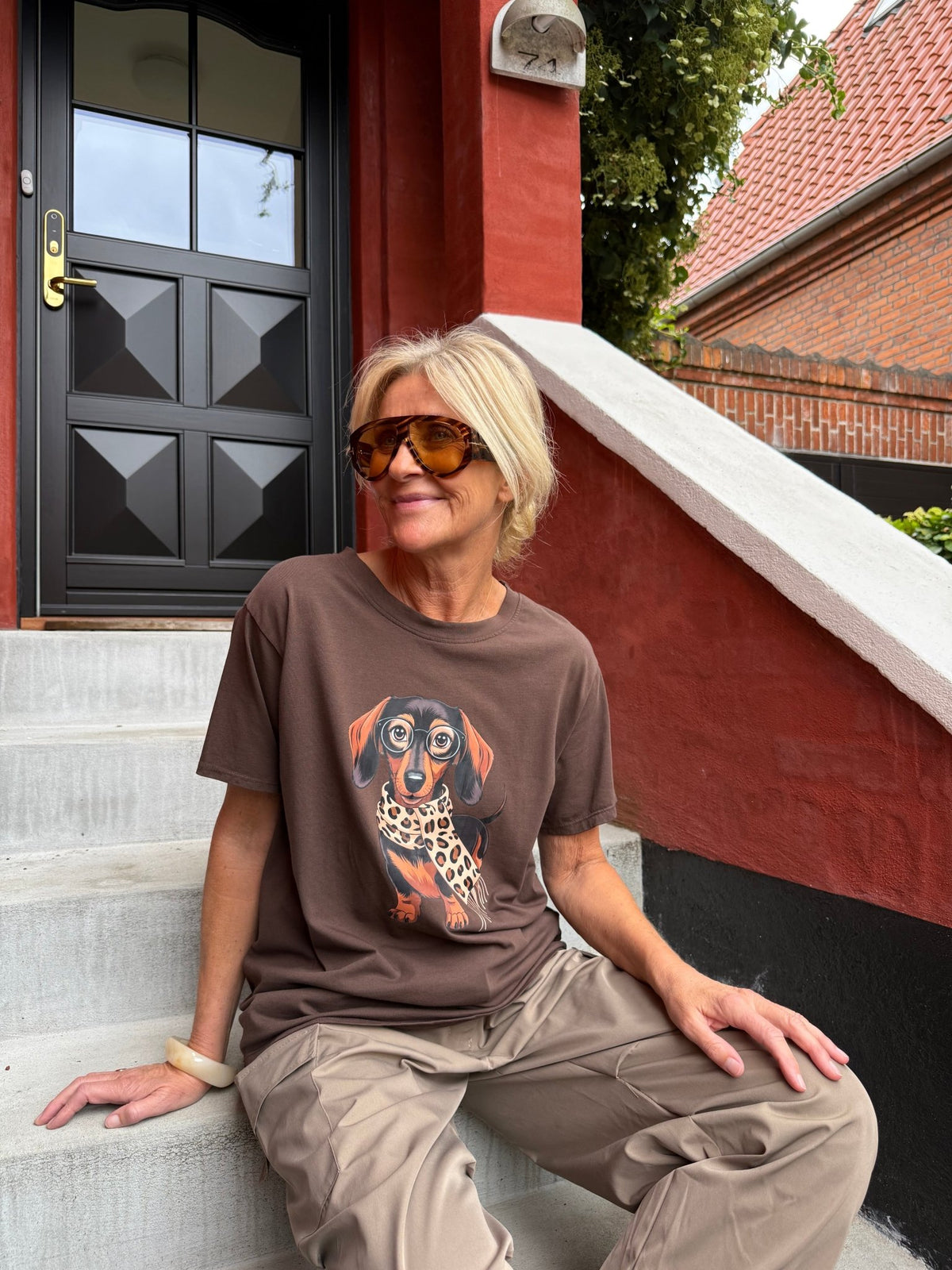 En kvinde iført en Inge Kakao-T-shirt fra Marta du Château og solbriller sidder på trappen uden for en rød murstensbygning.