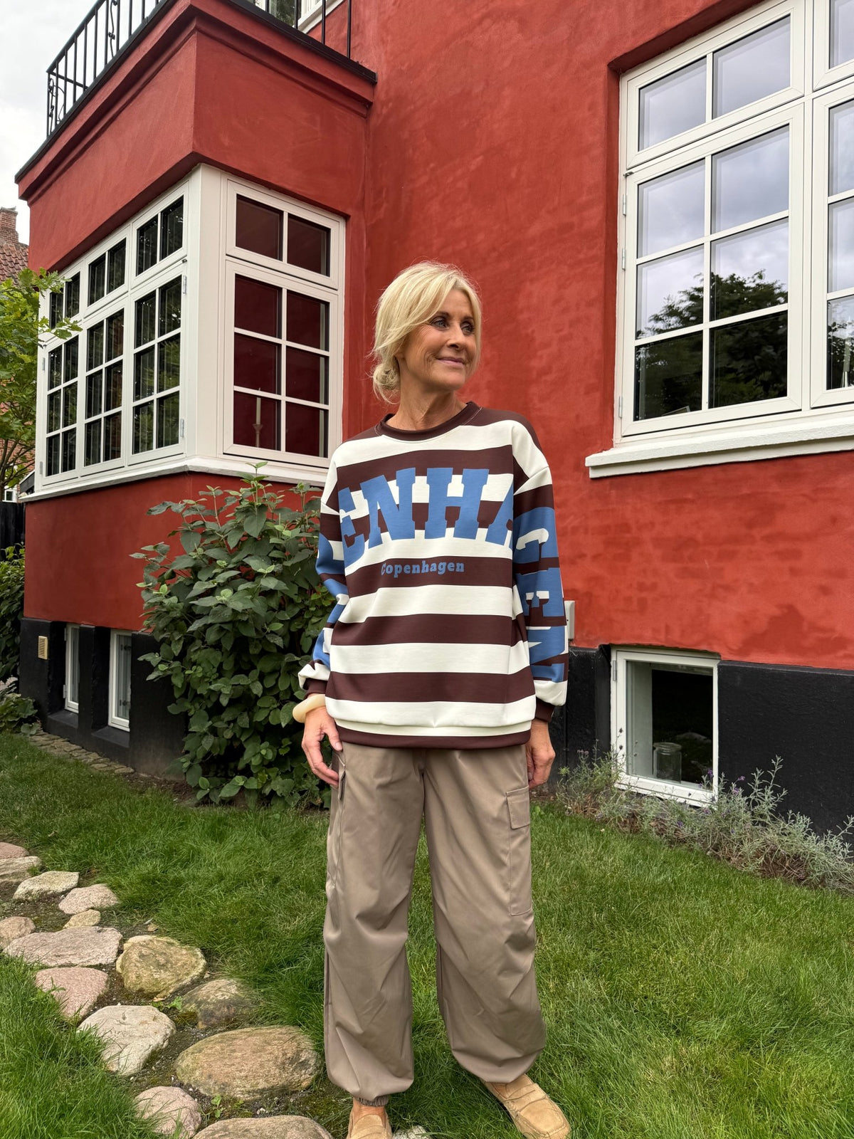 En kvinde i en sweatshirt med mønsteret »Marta du Château Nerina Brun« står uden for et rødt hus med store vinduer og grønne planter.