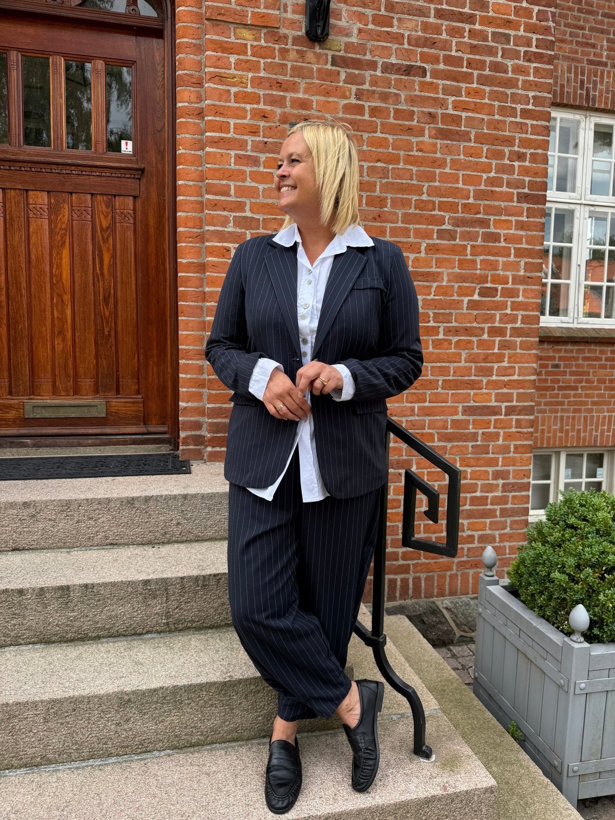 En person iført Marta du Château Nebula Mørkeblå-bukser smiler på stentrapperne uden for en murstensbygning.