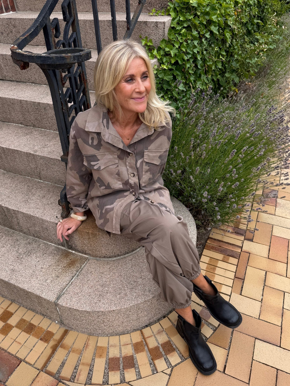 En kvinde med blondt hår i »Marta du Château – Moonstone Mønstret« og sorte støvler sidder på trappen og fremviser sin smarte stil.