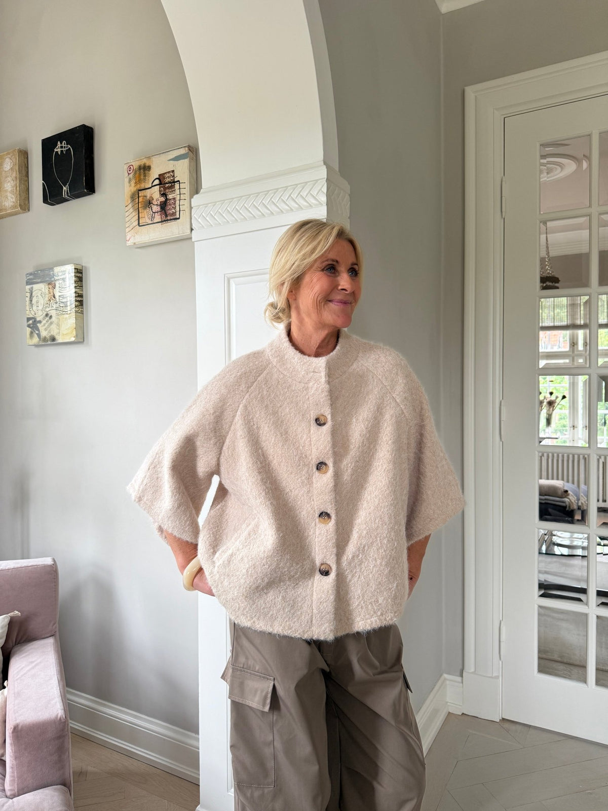 En kvinde står indendørs iført en Marta du Château Riviera Caramel-jakke, med kunst og et vindue i baggrunden.