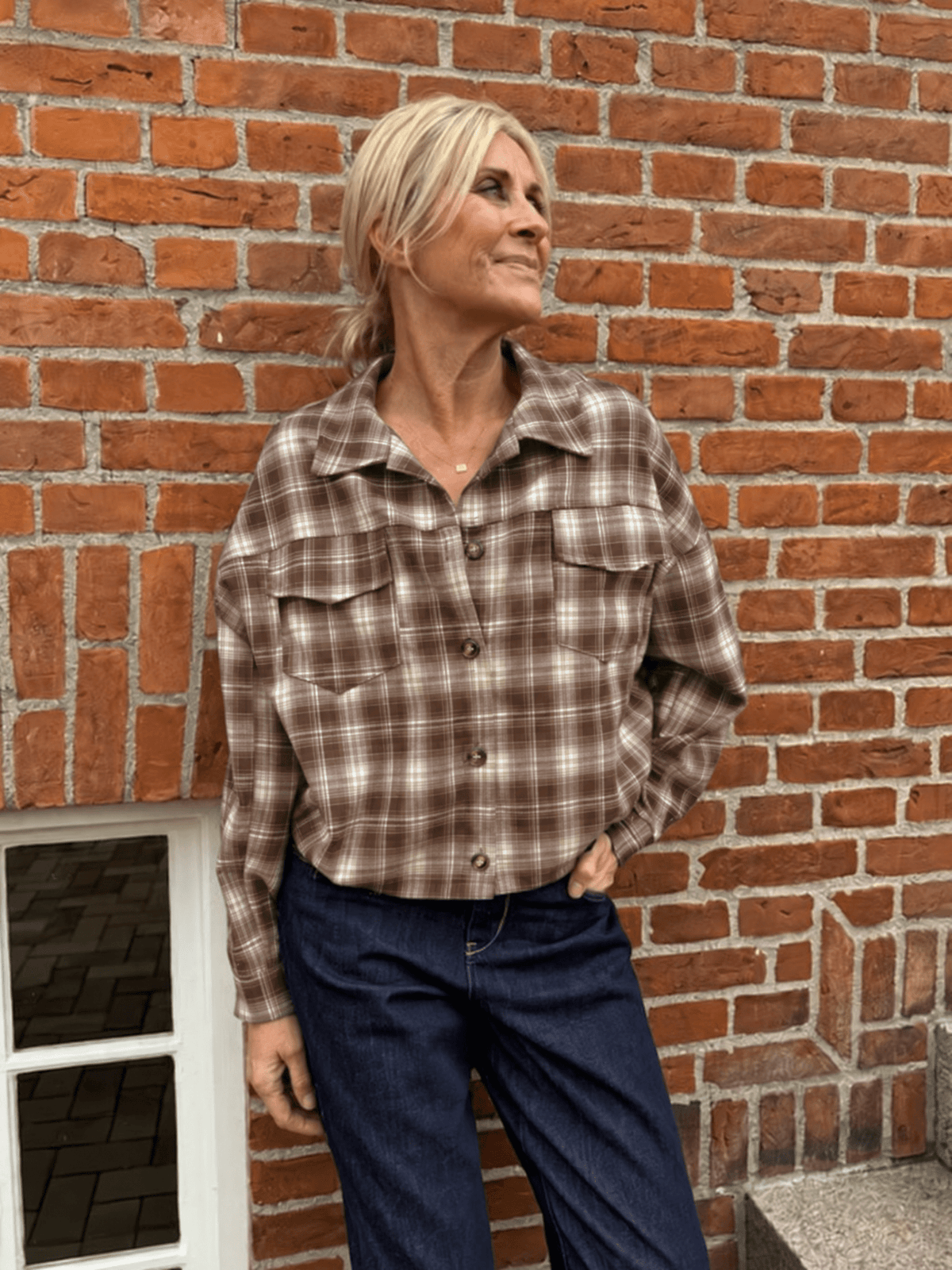 En kvinde iført en Marta du Château MdcMarbella Ternet-bluse og mørke jeans står ved en rød mur og ser tankefuld ud.
