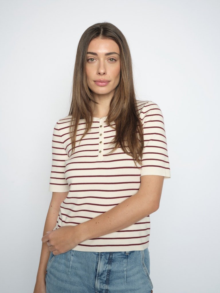 En kvinde iført en stribet bluse fra MOS MOSH Aliz Caliz og denimshorts står med ansigtet vendt mod kameraet foran en ensfarvet baggrund.